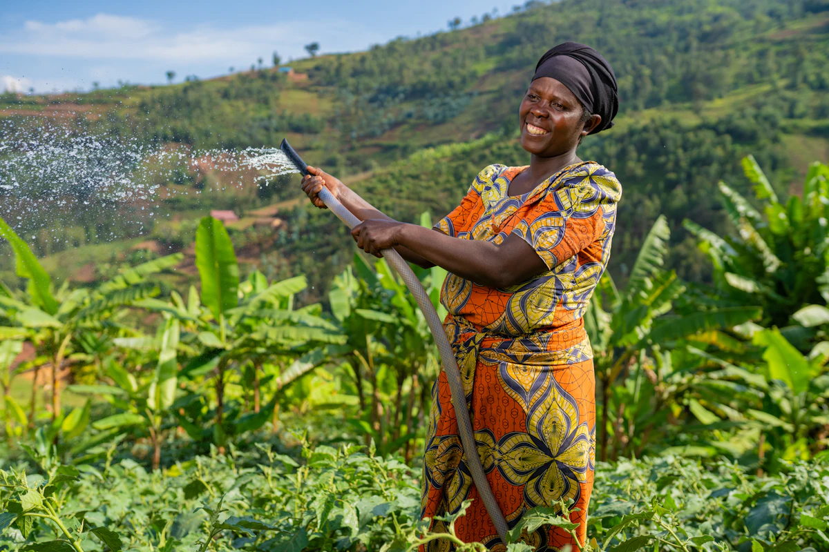 A Rwandan farmer in a bright orange dress smiles while watering her field with an irrigation hose.