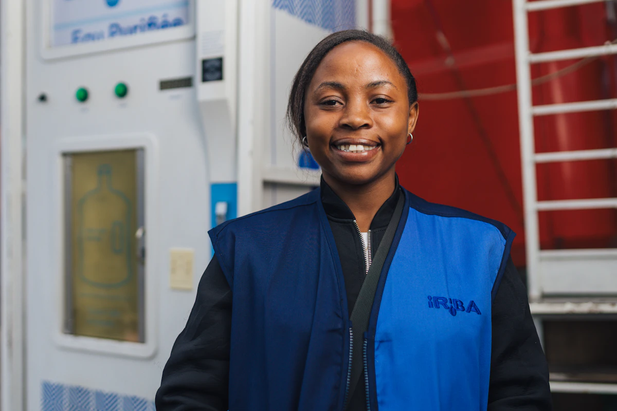 An IRIBA station operator in a blue vest smiles in front of a Tap&Drink machine.