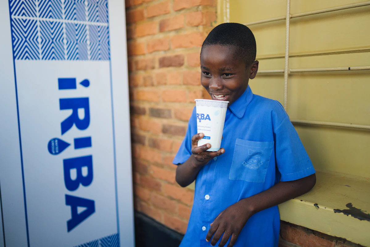 A schoolboy in a blue uniform smiles while holding an IRIBA paper cup outside a brick school building.
