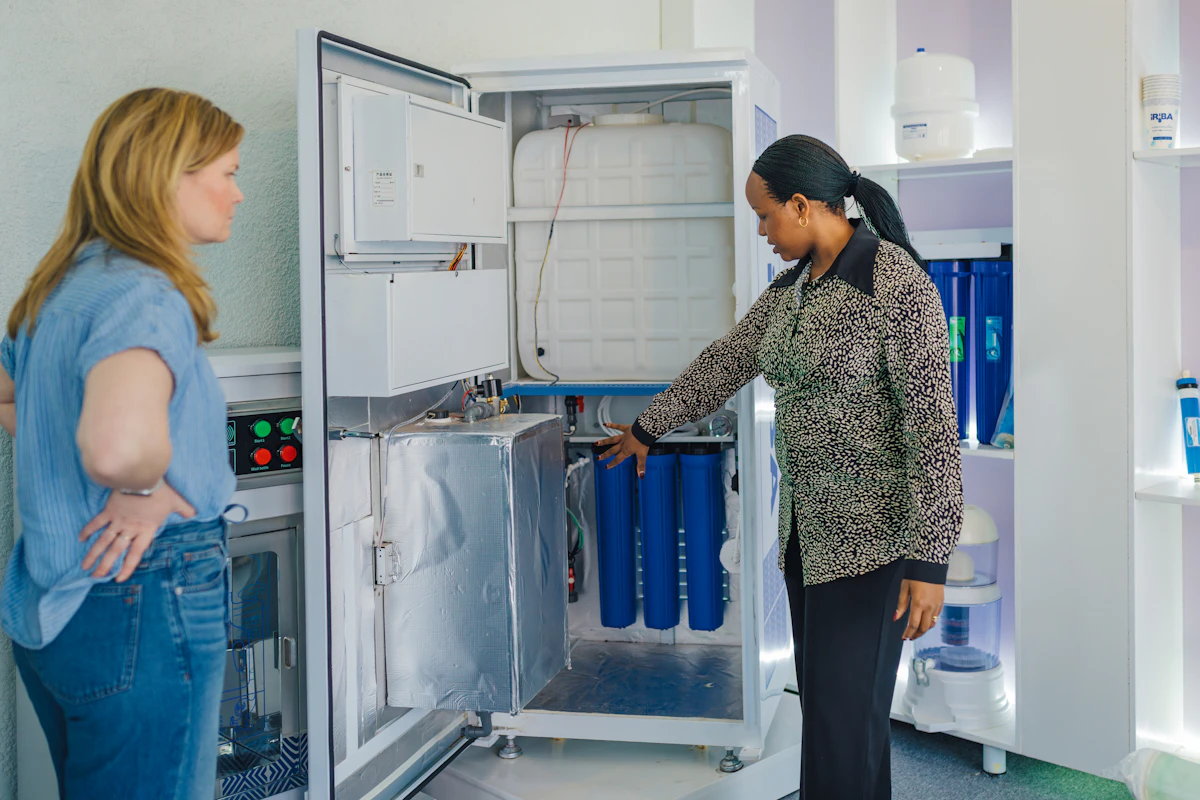 Two women examine the open interior of an IRIBA machine, showing its blue filter cartridges and tank.