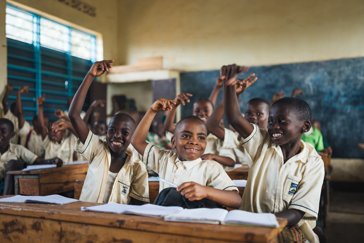 Smiling schoolchildren in matching cream uniforms sit at wooden desks in a classroom, eagerly raising their hands, with open notebooks in front of them and a chalkboard behind.