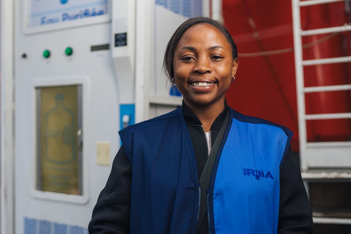 An IRIBA station operator in a blue vest smiles in front of a Tap&Drink machine.