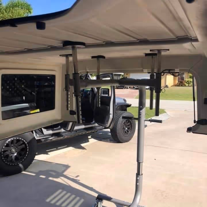 A Jeep with open doors parked on a driveway, viewed from inside a canopy or trailer with a mounted metal rack structure visible in the foreground.