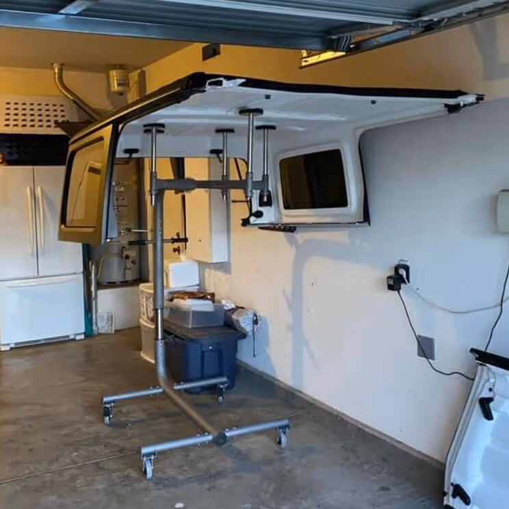 White vehicle camper shell mounted on a metal cart with wheels inside a garage with concrete floor and white walls.