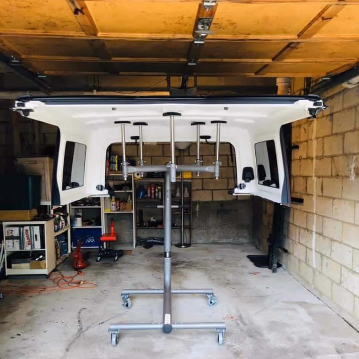 White truck bed shell supported on a wheeled adjustable metal stand inside a garage with shelves and tools in the background.