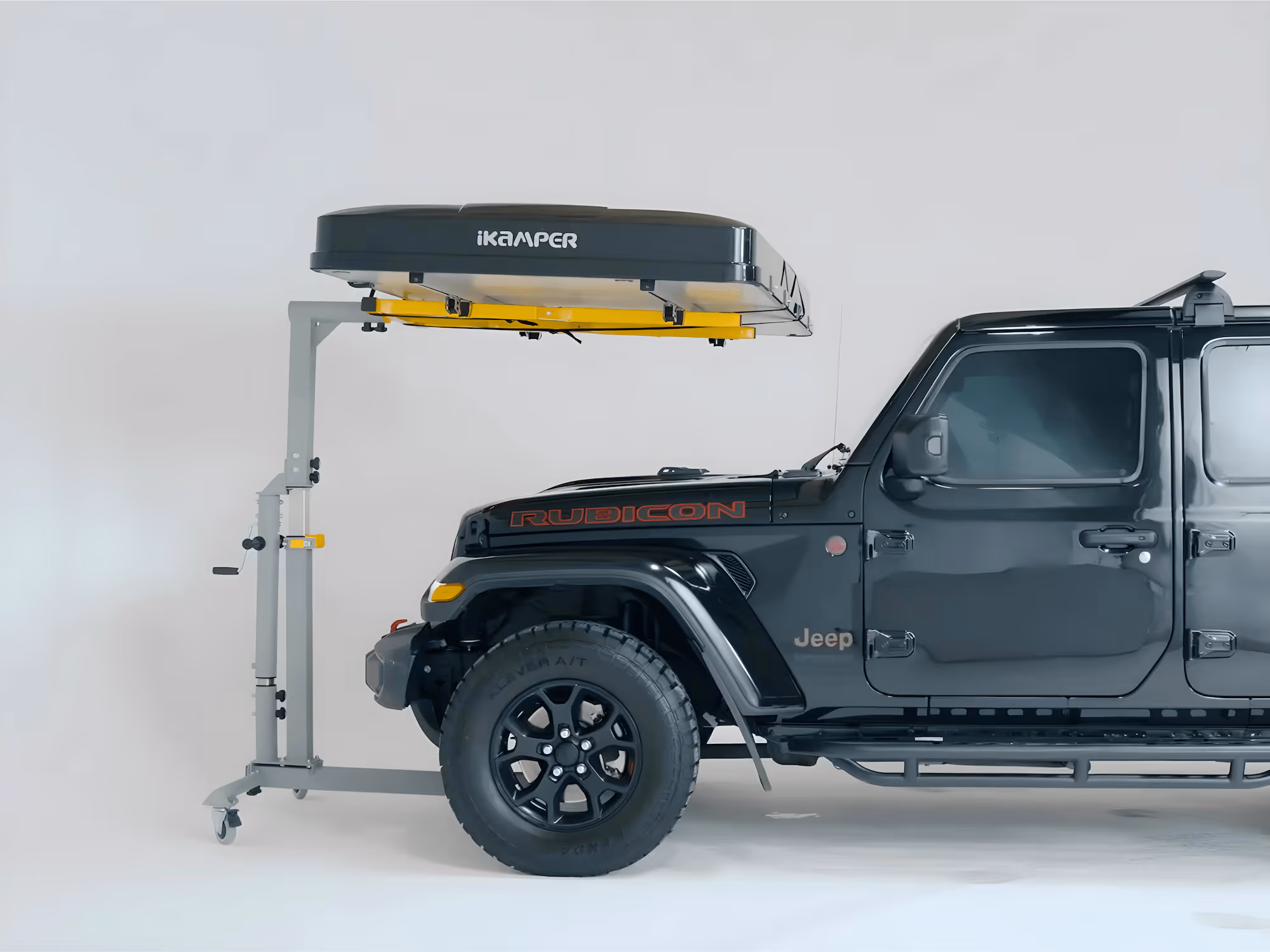 Black Jeep Rubicon parked indoors with an iKamper rooftop tent mounted on a gray stand beside it.