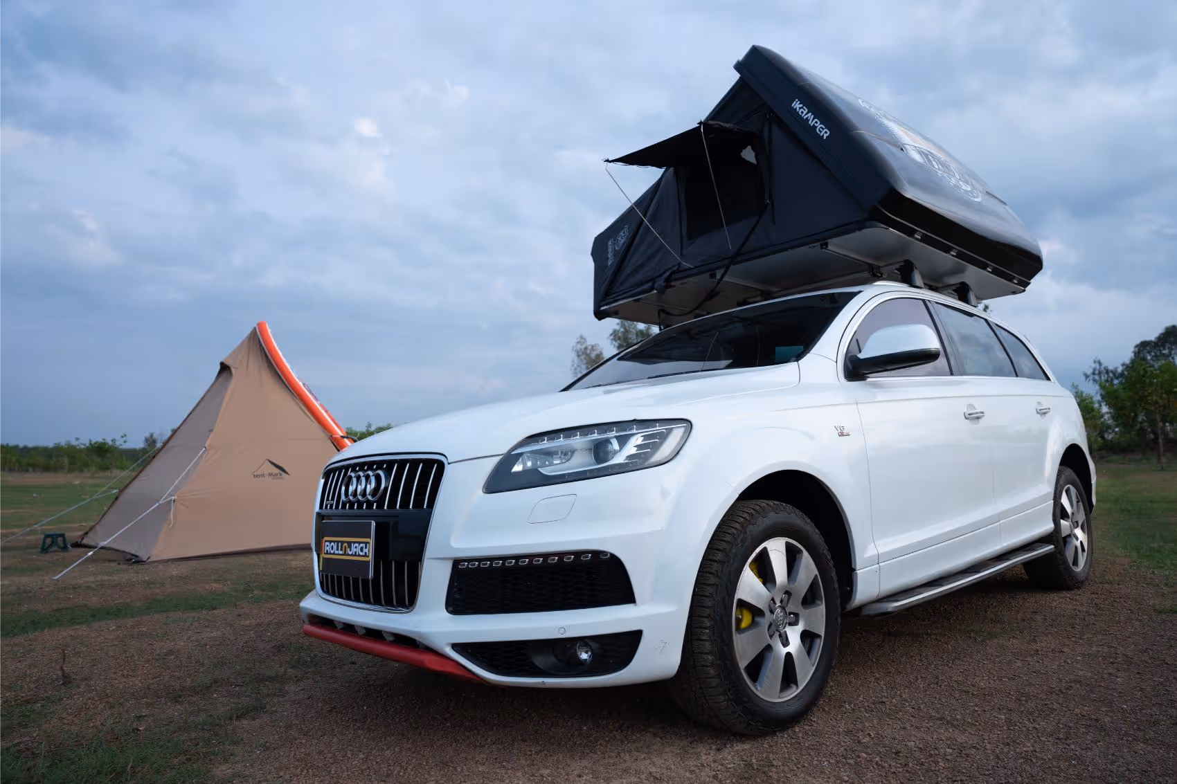 White Audi SUV parked on grass with a black roof tent deployed and a beige triangular camping tent in the background.