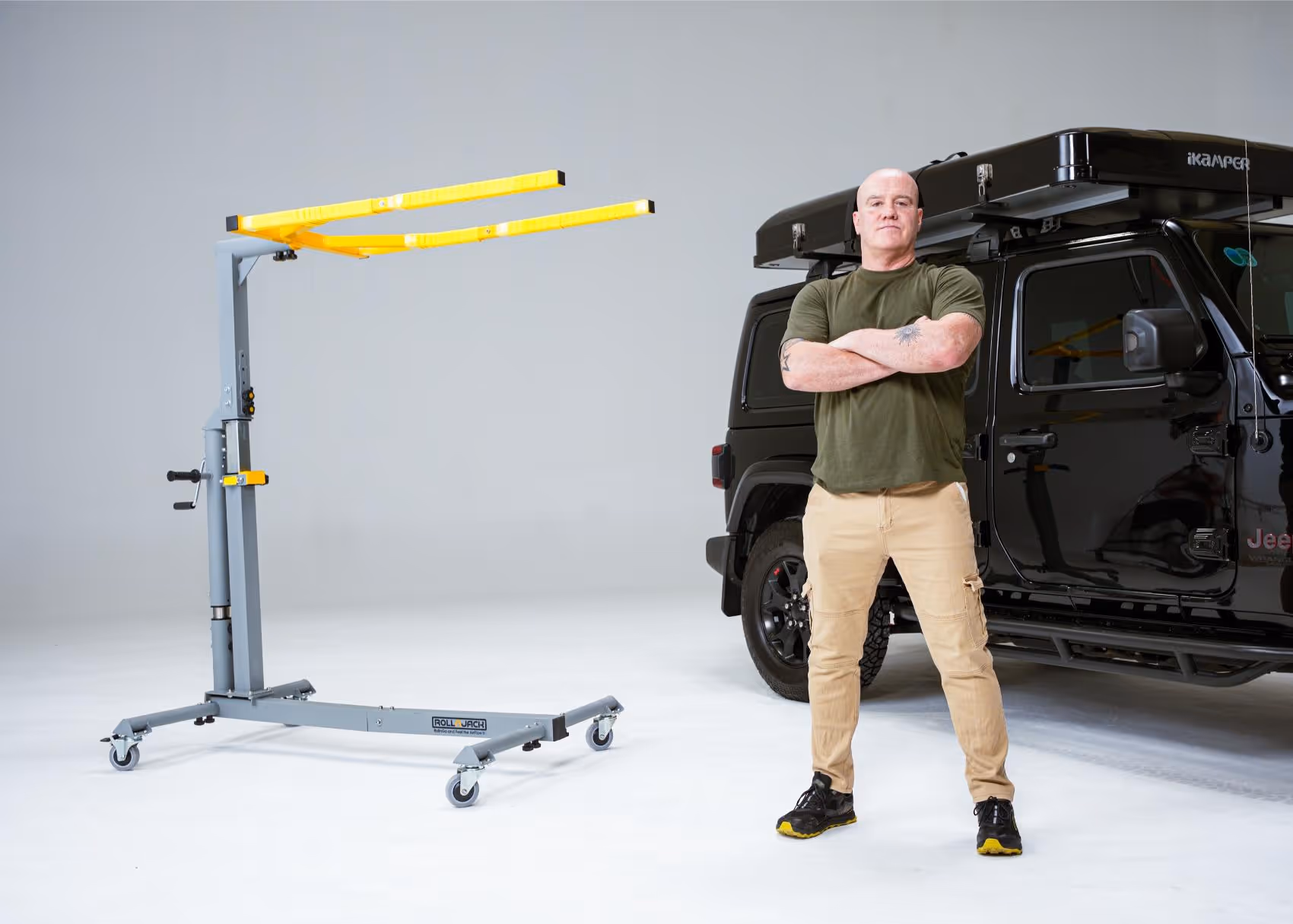 Man standing with crossed arms in front of a black Jeep with a roof tent and a gray and yellow Roll Jack tent lift next to him.