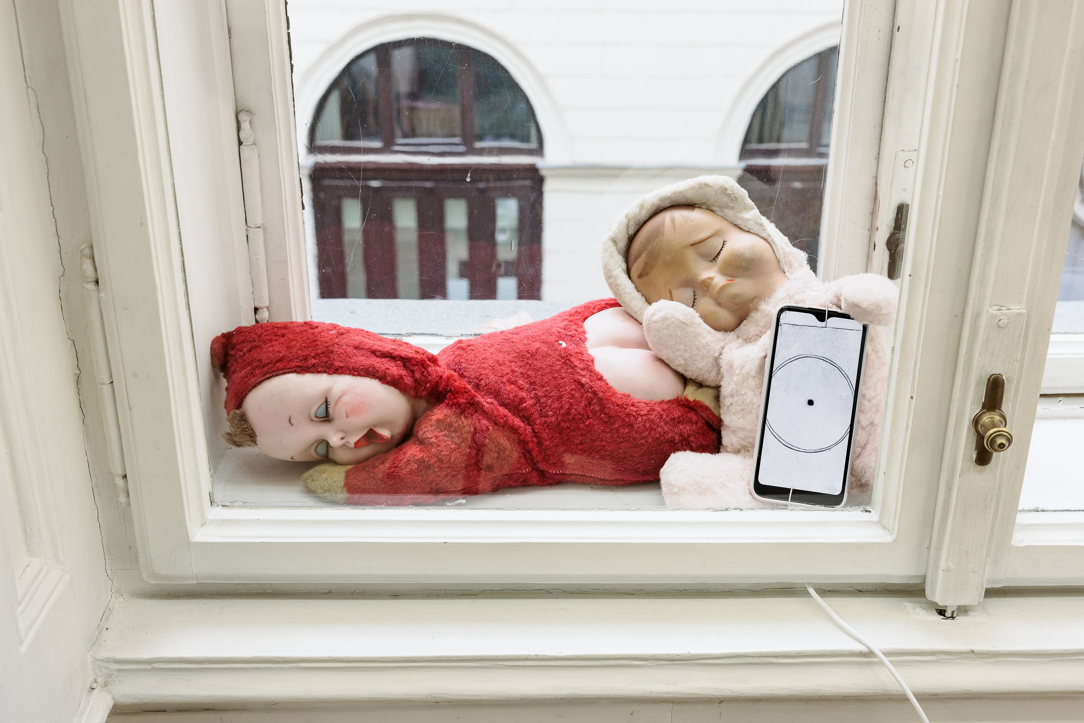 Nicole-Antonia Spagnola, Old Babies (Booming), 2026, Dolls, smartphone, USB-C charger, Mp4 (B/W), 60 × 23 × 23 cm (23 5/8 × 9 × 9 in). Courtesy of the artist and Felix Gaudlitz.