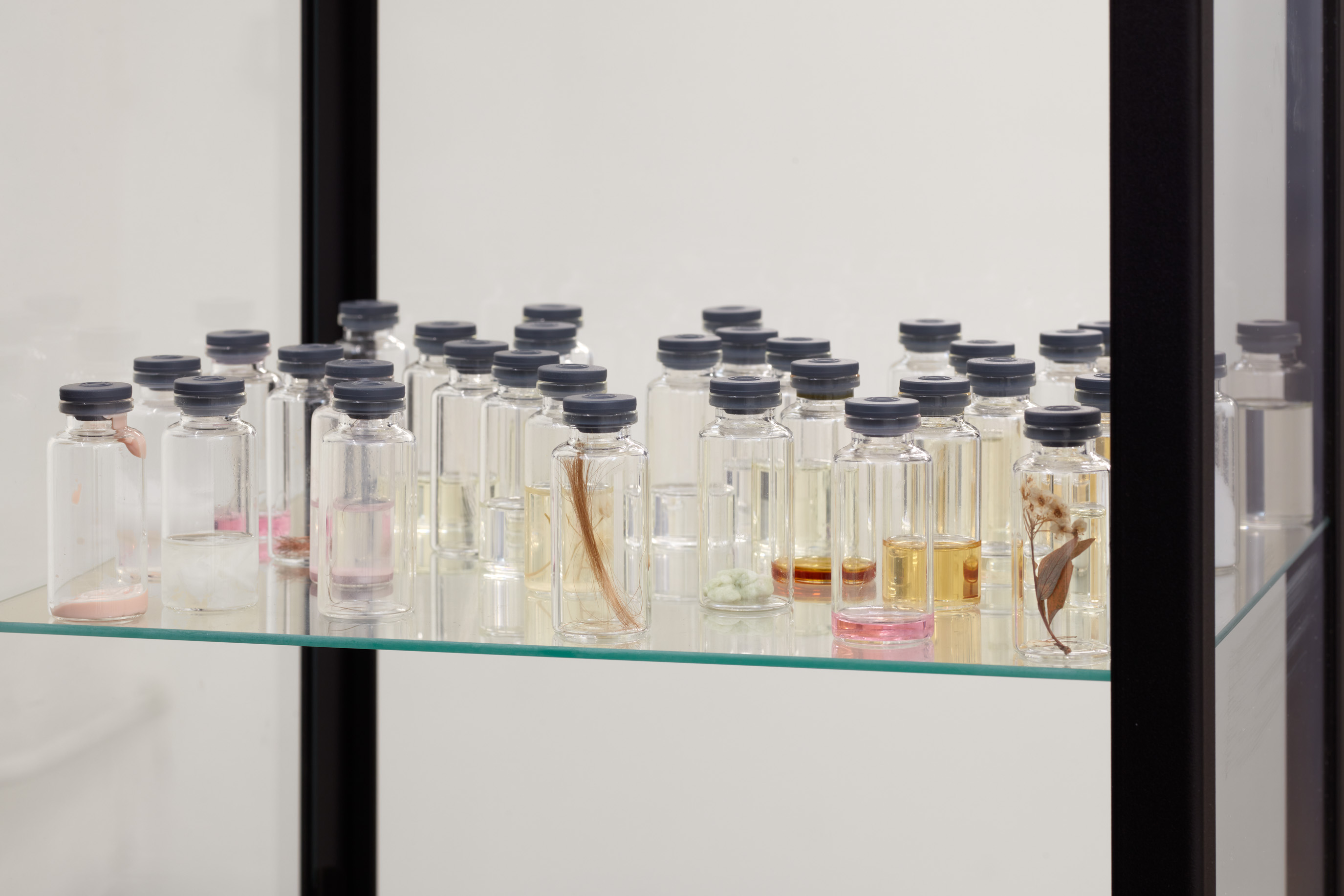 Detail. Ella Fleck, a zombie resurrected by a ghost, 2026. Glass cabinet, 100 borosilicate glass vials filled with Acetone (C4H8O), hair (keratin, α-helix polypeptide chains), saliva (aqueous mucin suspension, pH 6.2–7.6), urine-saturated cellulose substrate, chlorhexidine gluconate (C22H30Cl2N10 · 2C6H12O7), coffea arabica aqueous extract, prunus amygdalus dulcis oil, androstenone (C19H28O), acetic acid (CH3COOH), masticated polyisobutylene, mica (KAl2(AlSi3O10)(OH)2), MAC Retro Matte Lipstick (hydrogenated polyisobutene, pentaerythrityl tetra-di-t-butyl hydroxyhydrocinnamate, CI 15850, CI 77491), eyebrow hair (vellus keratin filament), polyethylene terephthalate fibre (PET, C10H8O4)n, galaxolide 50 (1,3,4,6,7,8- hexahydro-4,6,6,7,8,8-hexamethylcyclopenta[g]-2-benzopyran, C18H26O, in isopropyl myristate carrier), γ-octalactone (C8H14O2), santalum album oil, velvione (15-pentadecanolide, C15H28O2), α-ionone (C13H20O), methyl salicylate (C8H8O3), cadaverine (pentane-1,5- diamine, C5H14N2), hedione (methyl dihydrojasmonate, C13H22O3), pogostemon cablin extract, citrus sinensis essence, Wild rose hand cream (aqua, glycerin, helianthus annuus seed oil, rosa canina fruit oil, parfum), Baby powder fragrance oil (geraniol C10H18O, phenylethyl alcohol C8H10O, lilial C14H20O, ethyl vanillin C9H10O3, coumarin C9H6O2, galaxolide C18H26O), Almond Milk & Honey fragrance oil (γ-undecalactone C11H20O2, methyl anthranilate C8H9NO2, phenylacetic acid C8H8O2, benzyl benzoate C14H12O2, ethyl maltol C7H8O3, coumarin C9H6O2, vanillin C8H8O3), White Musk fragrance oil (iso E super C16H24O, habanolide C16H28O2, ambroxan C16H28O, benzyl acetate C9H10O2, methyl dihydrojasmonate C13H22O3, coumarin C9H6O2, indole C8H7N), Sweet Brazilian fragrance oil (ethyl vanillin C9H10O3, γ-nonalactone C9H16O2, coumarin C9H6O2, tonalide C18H26O, δ-decalactone C10H18O2, benzyl salicylate C14H12O3, iso E super C16H24O), dried Gypsophila, pressed Narcissi, dimensions variable; cabinet: 59.5 x 13.25 x 12 in. (151 x 33.5 x 30.5 cm). Image courtesy of Ehrlich Steinberg, Los Angeles. Photo: Evan Walsh.