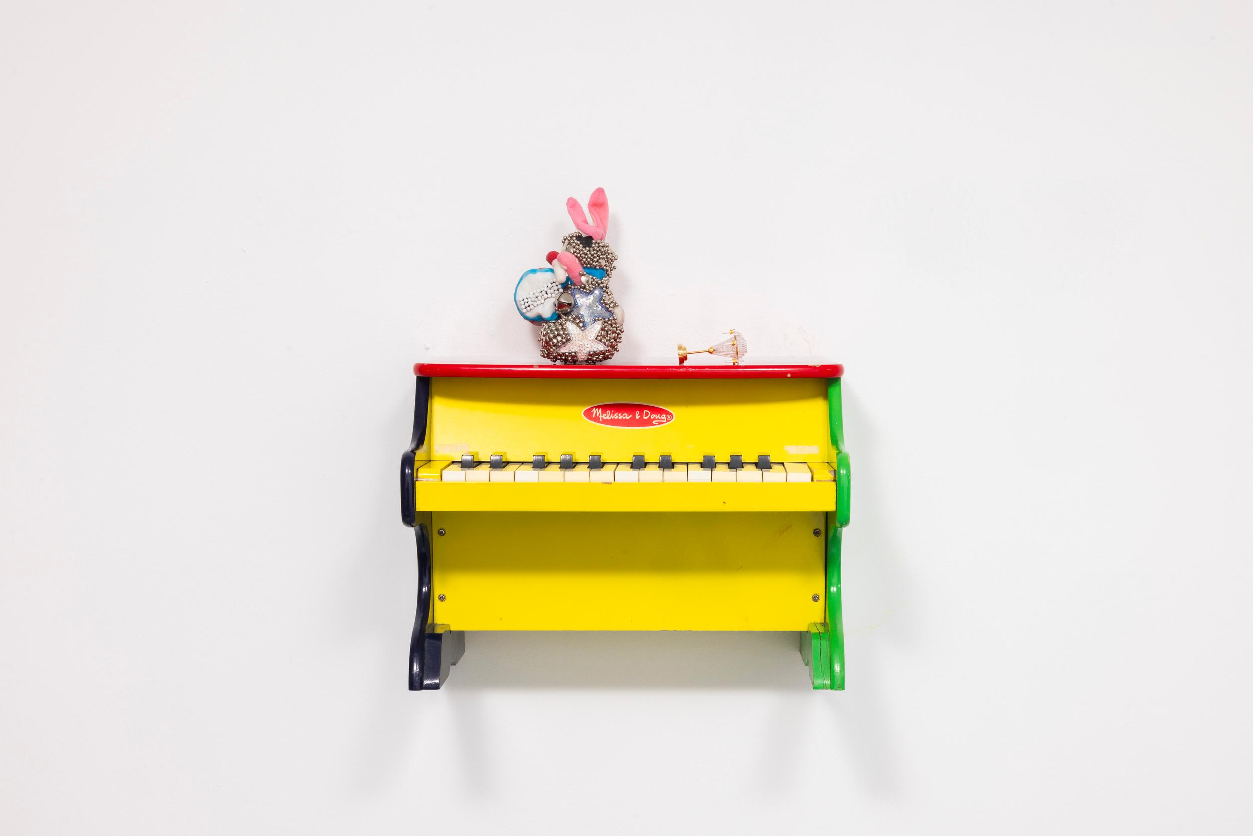 Faith Icecold, Act III, Scene I, 2026, Gift box bow, toy chandelier, adhesive, and vinyl on toy upright piano, Dimensions variable. Courtesy of the artist and Romance.