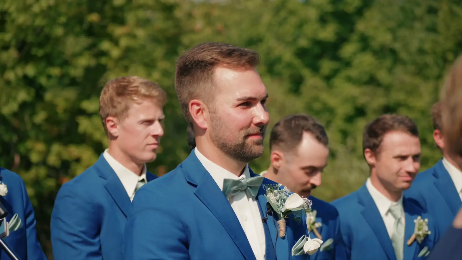Groom Joey stands with his groomsmen, wearing blue suits and white boutonnieres, during a sunny outdoor wedding at Ciccone Vineyard & Winery, Suttons Bay, Michigan.