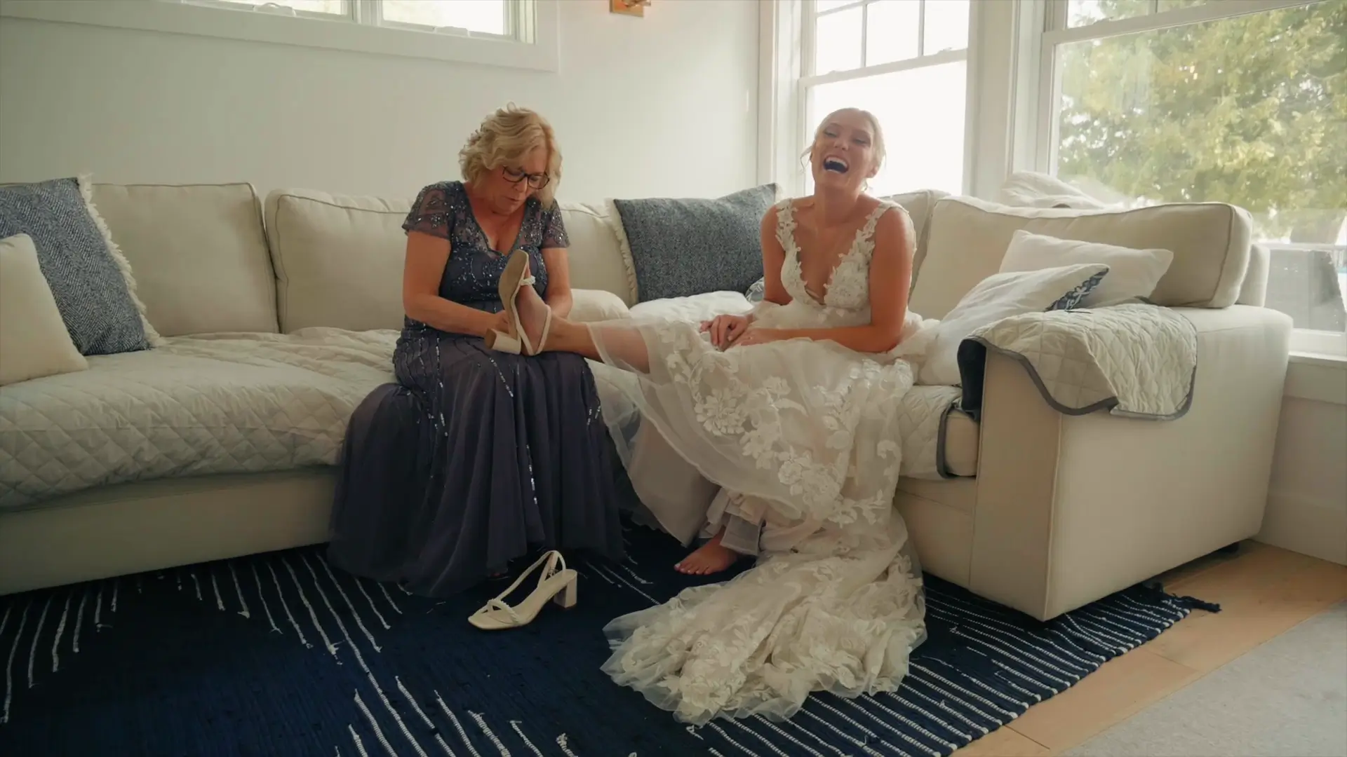 Bride Lindsey laughs on a couch as an older woman helps her put on a shoe. Lindsey is in a lace wedding dress, and there are pillows nearby. Natural light from large windows illuminates the cozy setting.