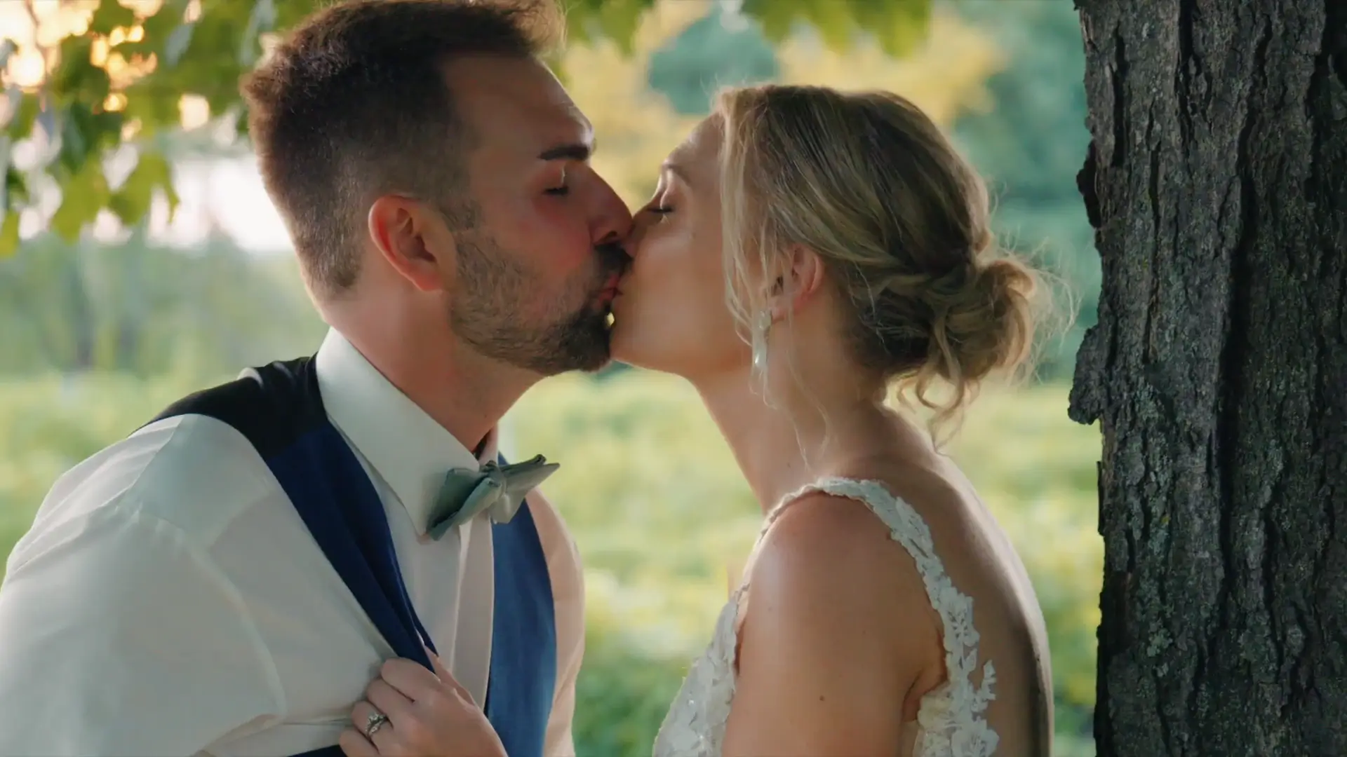 Couple sharing a tender kiss under a tree at Ciccone Vineyard & Winery in Suttons Bay, Michigan. The groom wears a suit with a bow tie, while the bride is in a lace gown. The sun-dappled greenery adds a romantic backdrop.