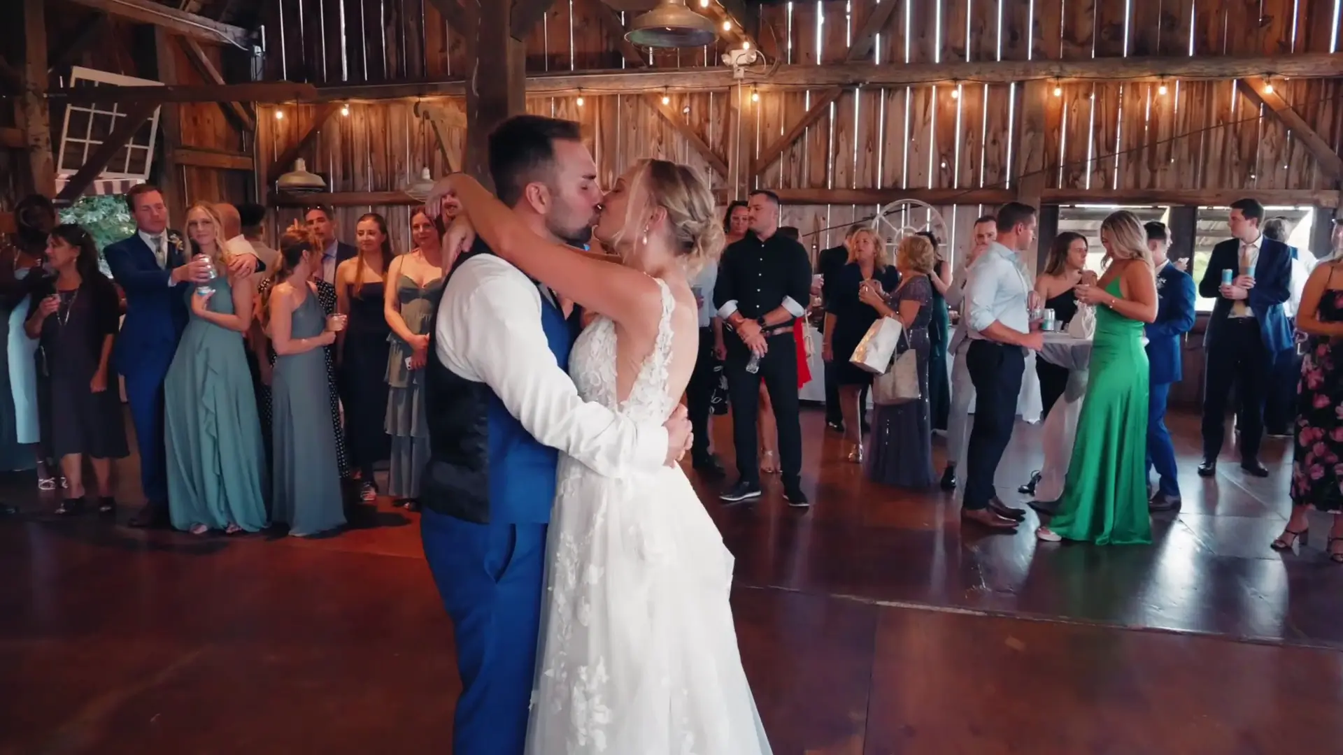 In this snapshot from their wedding highlight, Lindsey and Joey share a kiss on the dance floor at Ciccone Vineyard and Winery, surrounded by guests. The rustic Michigan barn setting adds charm to this joyful moment captured by their videographer.