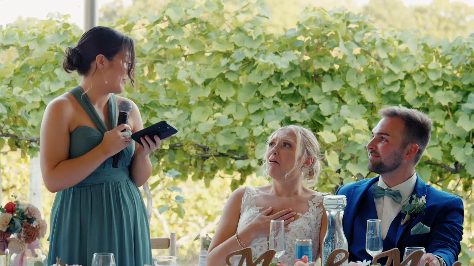 Bridesmaid in a green dress holds a microphone, making a speech to the bride and groom seated at a table. The bride looks touched, and the groom smiles. Greenery is visible in the background at Ciccone Vineyard and Winery.