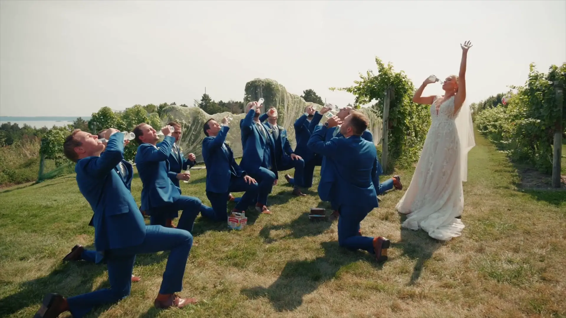 Bride and groomsmen, in blue suits, celebrate in a vineyard, joyfully holding drinks amidst lush Michigan greenery at Ciccone Vineyard and Winery. The bride raises her arm triumphantly, capturing a moment of fun and laughter on their special day.