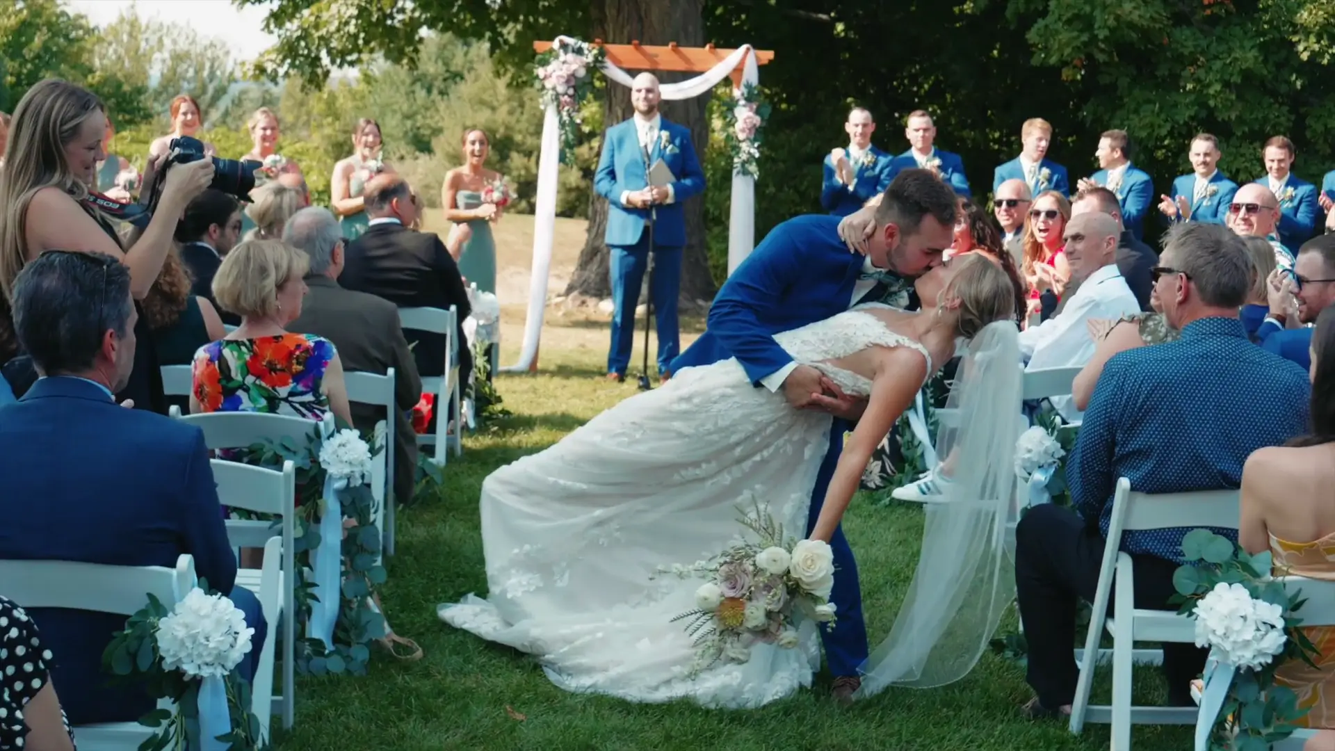 Bride and groom share a romantic kiss during their wedding ceremony at Ciccone Vineyard & Winery, Suttons Bay. Surrounded by guests and lush greenery, the bride holds a bouquet as the groom dips her.