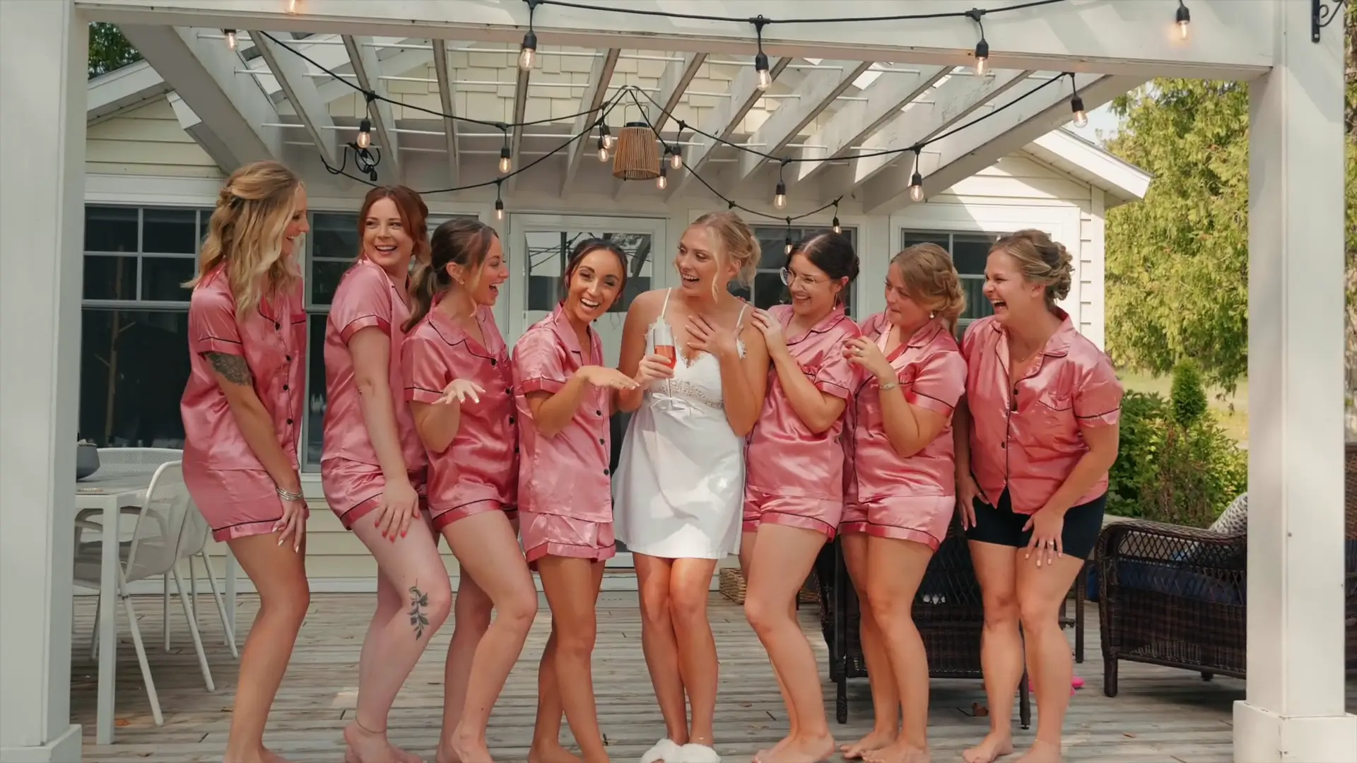 Bride Lindsey in a white robe is surrounded by her bridesmaids in matching pink pajamas, all smiling and laughing under a pergola with string lights at Lindsey's AirBnB.