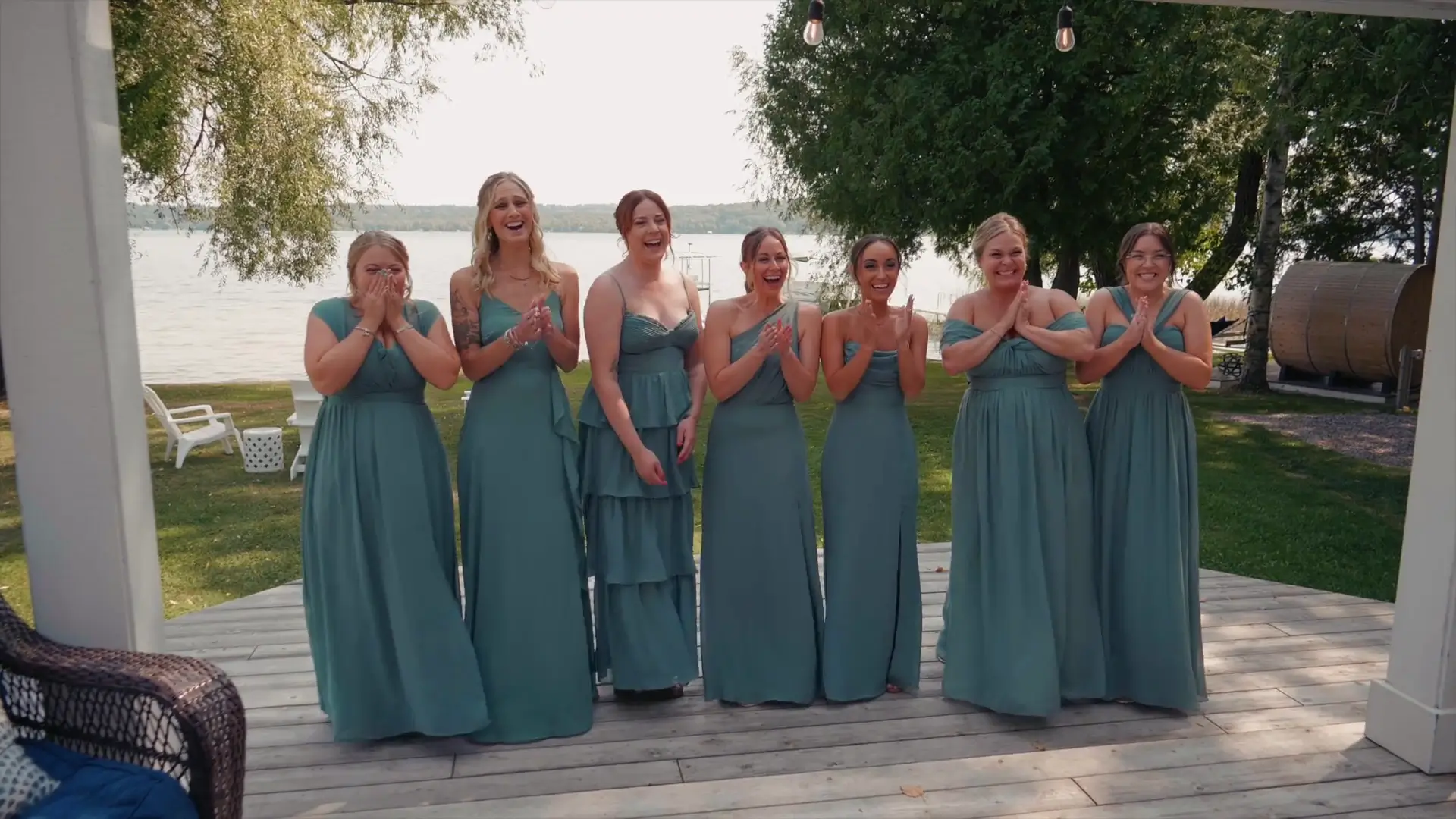 Eight bridesmaids in teal dresses smiling and clapping joyfully in front of a serene Michigan lake backdrop.