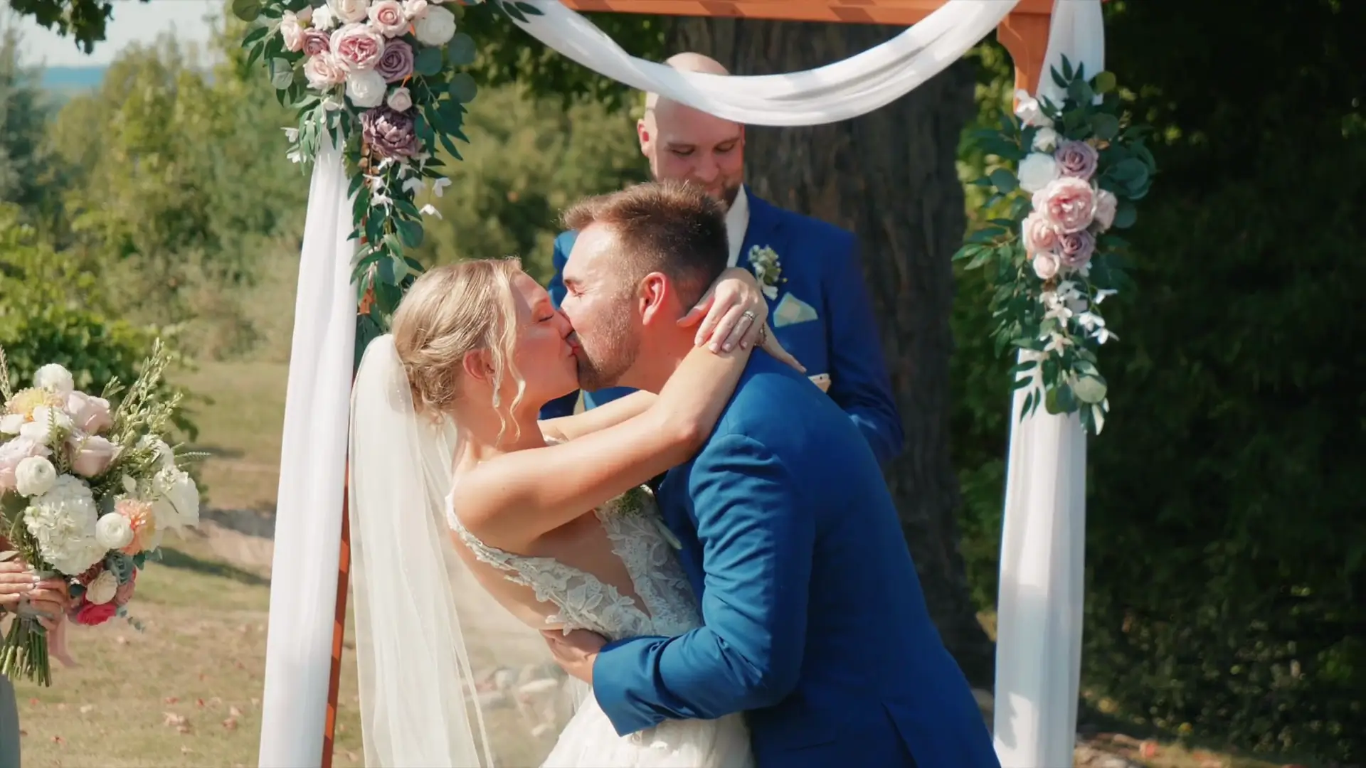 In this still from their wedding video, Lindsey and Joey share a kiss under a floral arch at Ciccone Vineyard and Winery. The lush, Michigan greenery adds a vibrant backdrop to this special moment captured by their wedding videographer.