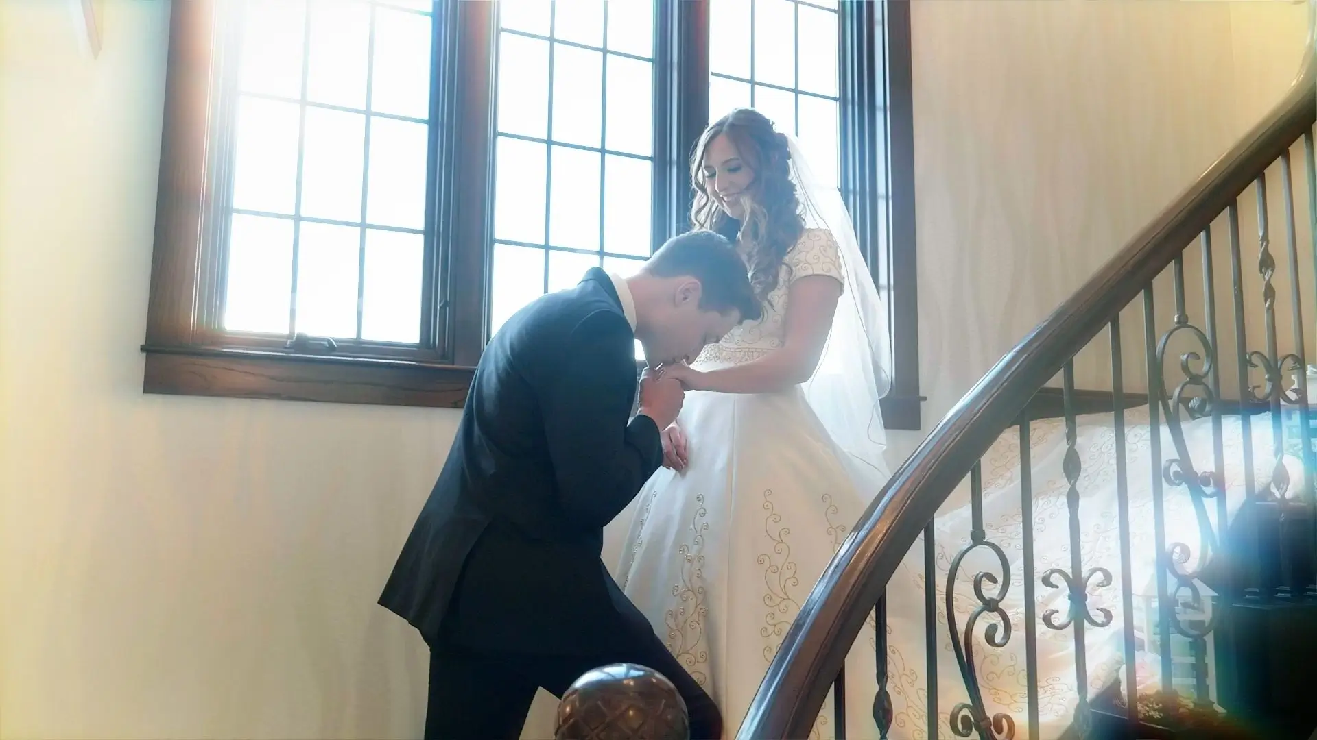Bride Maddie and groom Jack share a tender moment on the ornate staircase at Venue3Two, Grand Rapids, Michigan. Maddie, in a vintage-inspired gown, smiles as Jack kneels and kisses her hand, bathed in soft natural light from tall windows.
