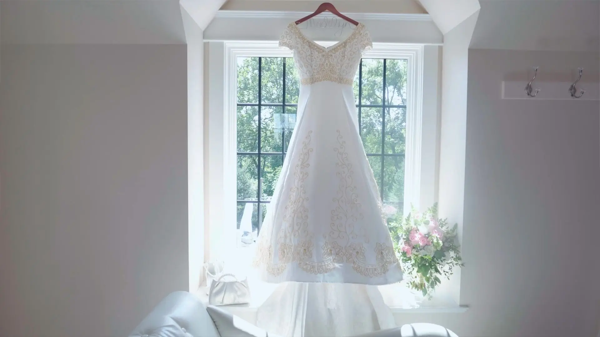 A beautiful wedding dress with intricate gold embroidery hangs in front of a bright window, adorned with a bouquet of pink and white flowers on the windowsill.
