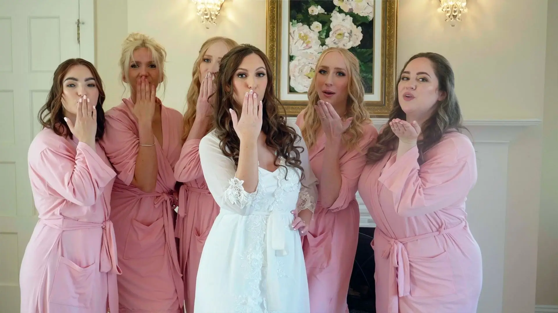 Bridal party in pink robes, with the bride in a white robe, blowing kisses toward the camera. They're standing in front of a framed floral painting at Venue3Two in Grand Rapids, Michigan.