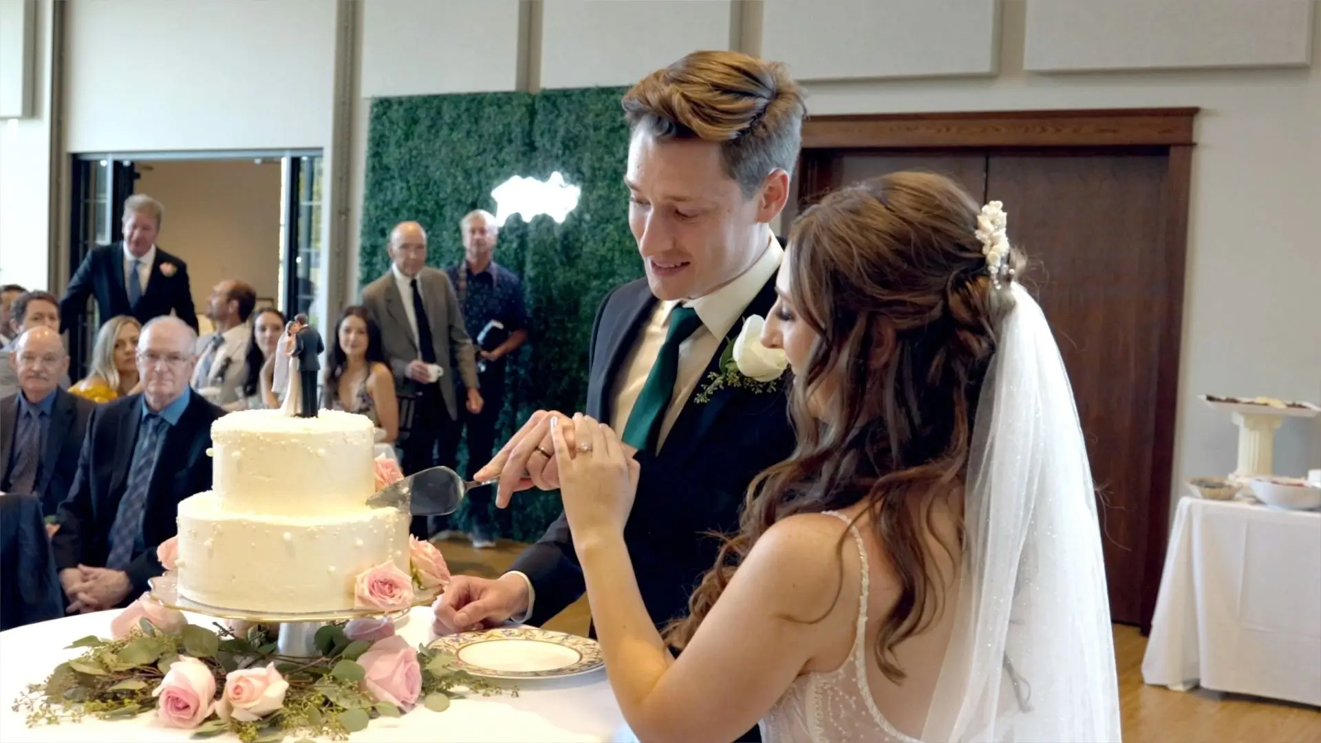 Maddie and Jack are cutting their wedding cake in front of guests at Venue3Two in Grand Rapids, Michigan. The cake is decorated with pink roses and a couple figurine topper. Guests are seated and watching the couple's special moment.
