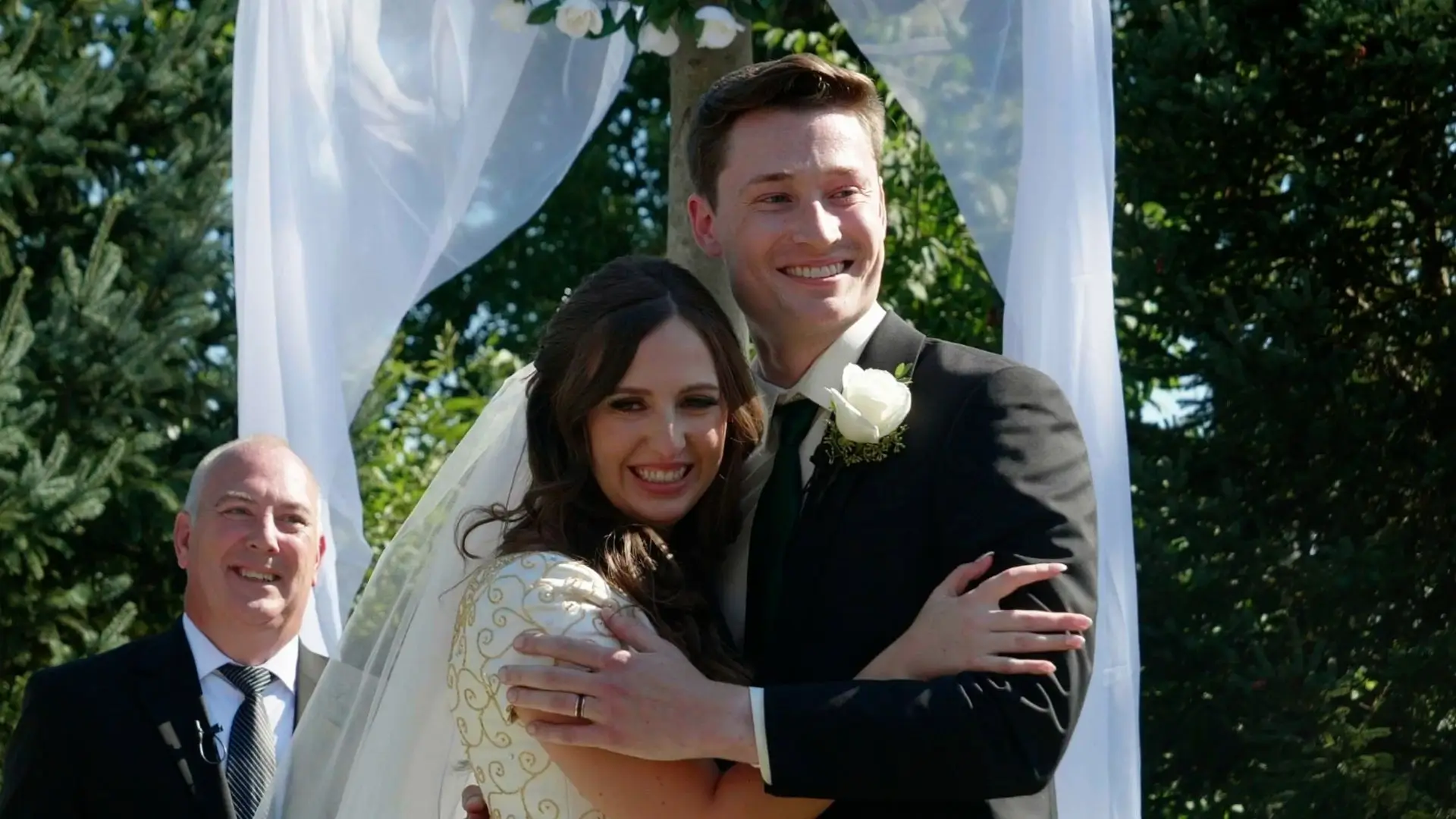 Bride Maddie and groom Jack embracing under an archway with lush Michigan greenery in the background, smiling happily at Venue3Two, Grand Rapids, Michigan.