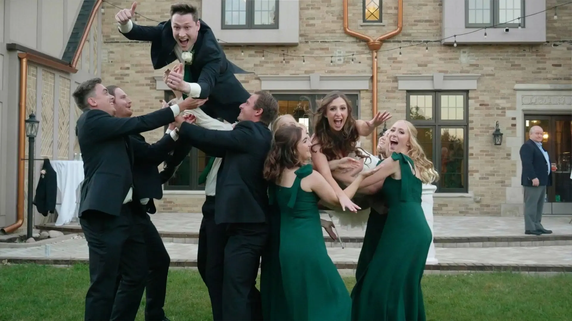 A joyful wedding moment at Venue3Two in Michigan, with groomsmen in suits tossing the groom and bridesmaids in green dresses tossing the bride and cheering, in front of the elegant brick venue.