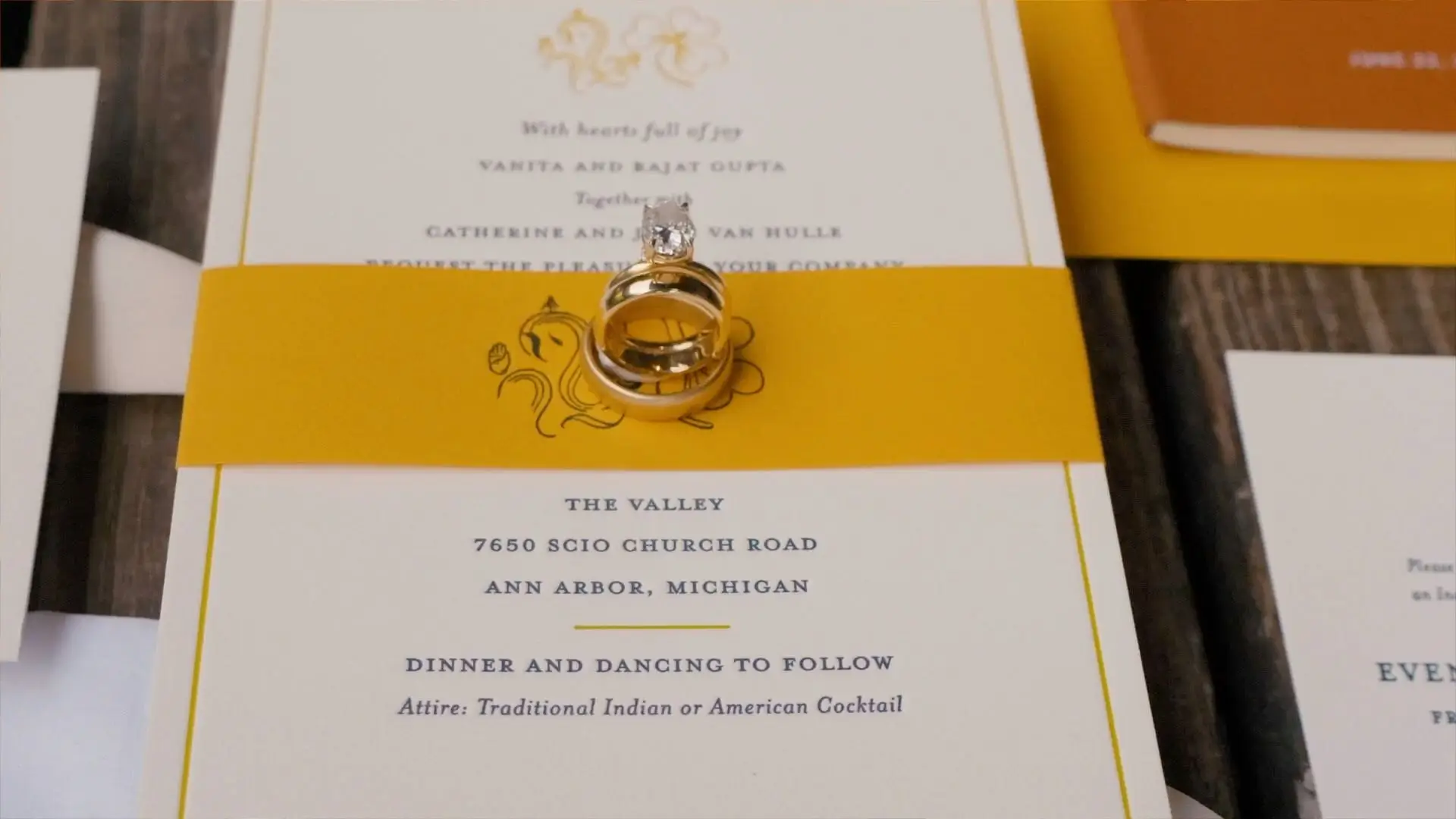 
Close-up of a wedding invitation for Anushka and Sean in Ann Arbor, Michigan. The invitation is white with a yellow band featuring an illustration of Lord Ganesha. Two gold rings with a diamond rest on top of the band.