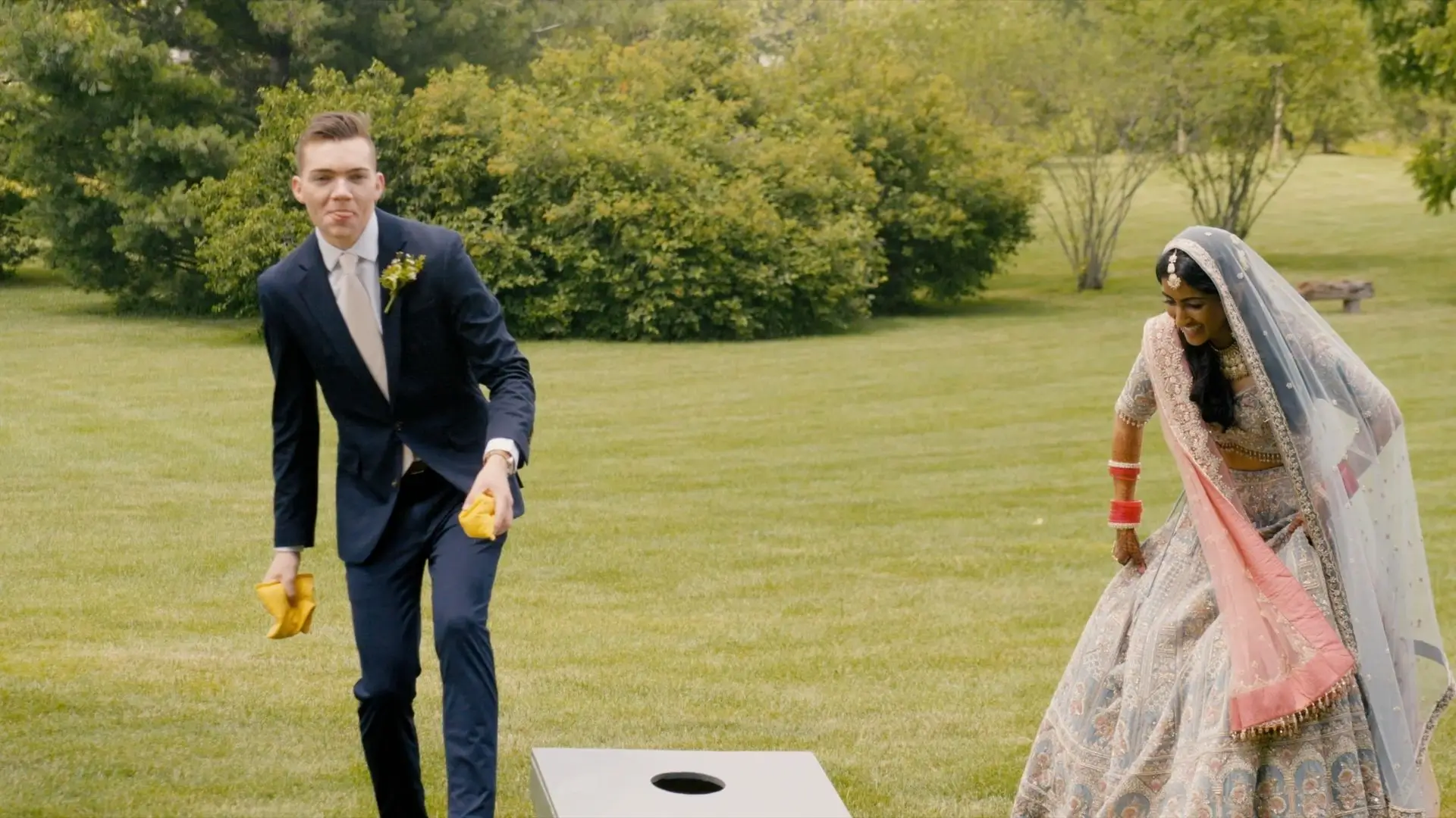 Bride Anushka dressed in a traditional Indian outfit and groom Sean in a western suit play cornhole outdoors at The Valley at Frutig Farms, Ann Arbor. Sean tosses a beanbag while Anushka smiles. Greenery is visible in the background.