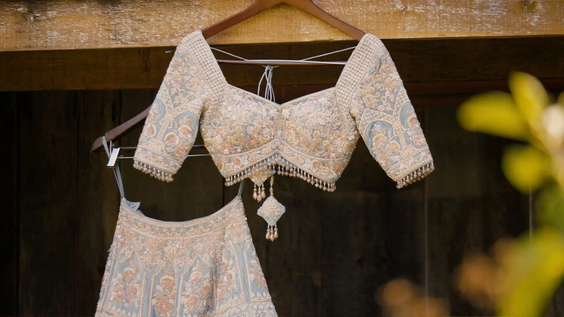 An intricately embroidered blue and gold bridal lehenga hangs from a wooden beam, with detailed beadwork and floral patterns, displayed in preparation for Anushka and Sean's wedding at The Valley at Frutig Farms in Ann Arbor, Michigan.