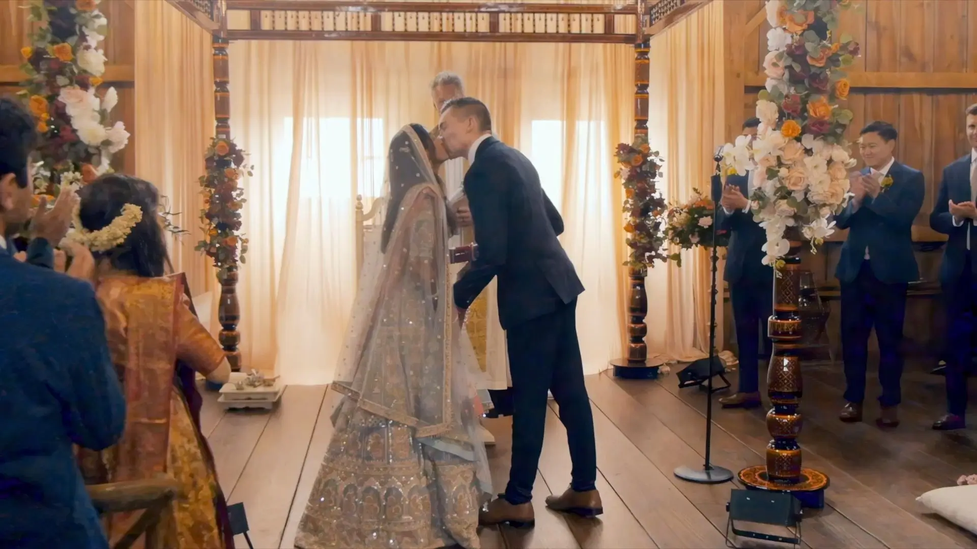Anushka and Sean share their first kiss during their wedding ceremony at The Valley at Frutig Farms in Ann Arbor, Michigan. The ceremony features vibrant floral decorations with guests clapping and celebrating in the background.