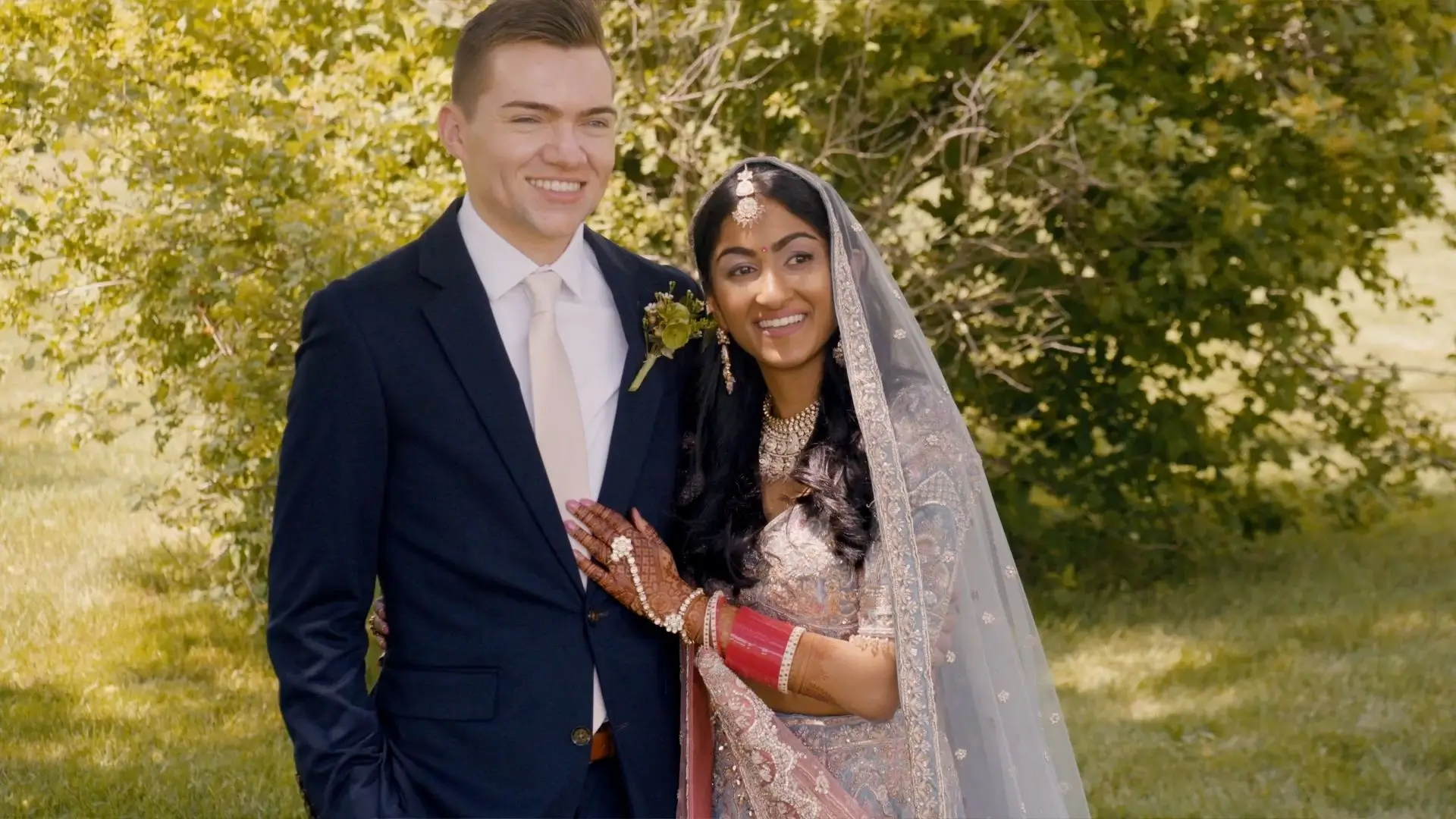Anushka and Sean, the bride and groom, stand outdoors at The Valley at Frutig Farms in Ann Arbor. Anushka wears a traditional bridal outfit with a veil and jewelry, while Sean is in a navy suit with a boutonniere. Both are smiling joyfully together.