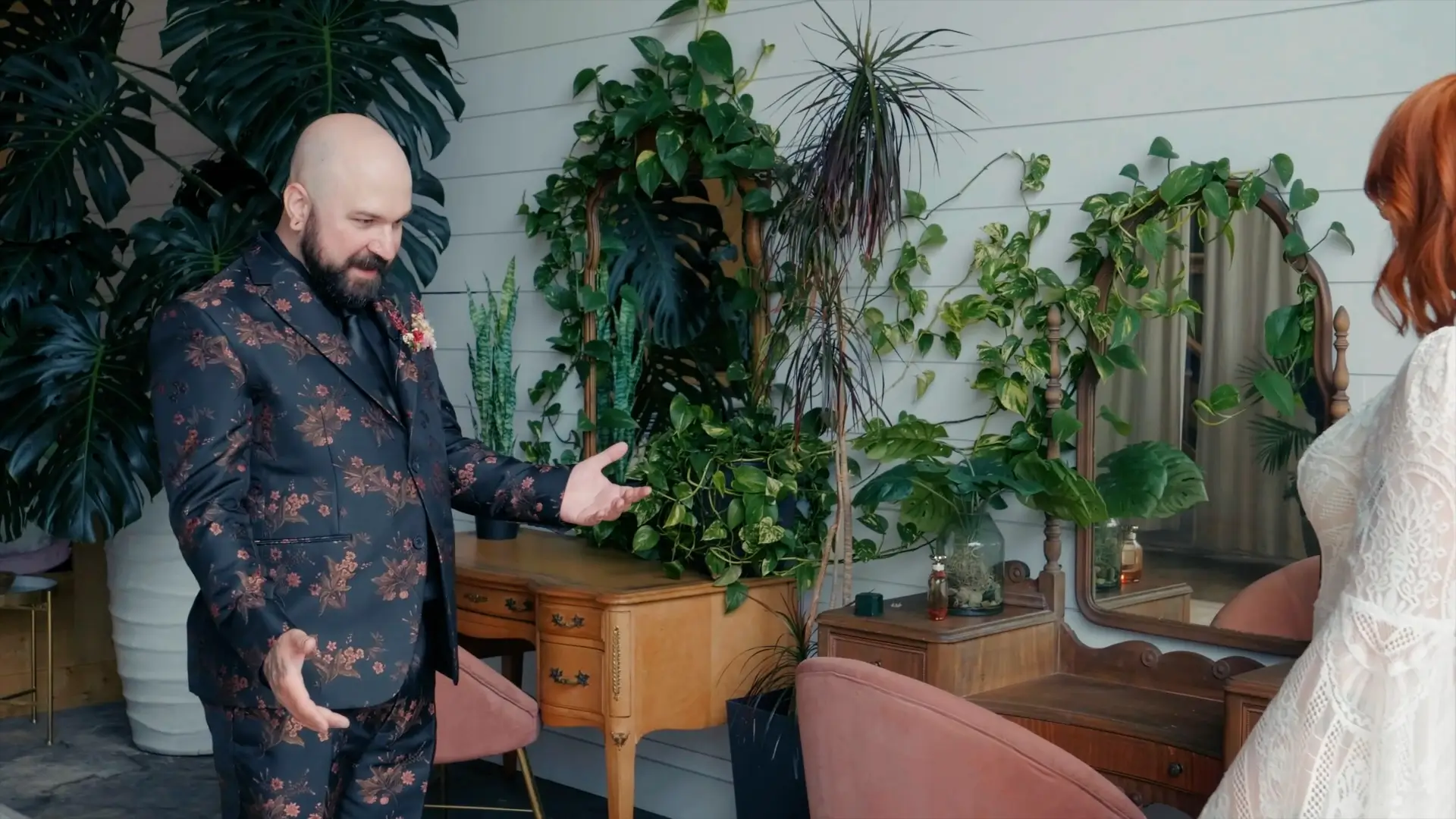 In this moment captured by the wedding videographer, the groom, dressed in a floral suit, gestures warmly in a plant-filled room, with a vintage vanity behind him, reflecting the hipster and artsy vibe of Kate and Zack's wedding in Stevensville, Michigan.
