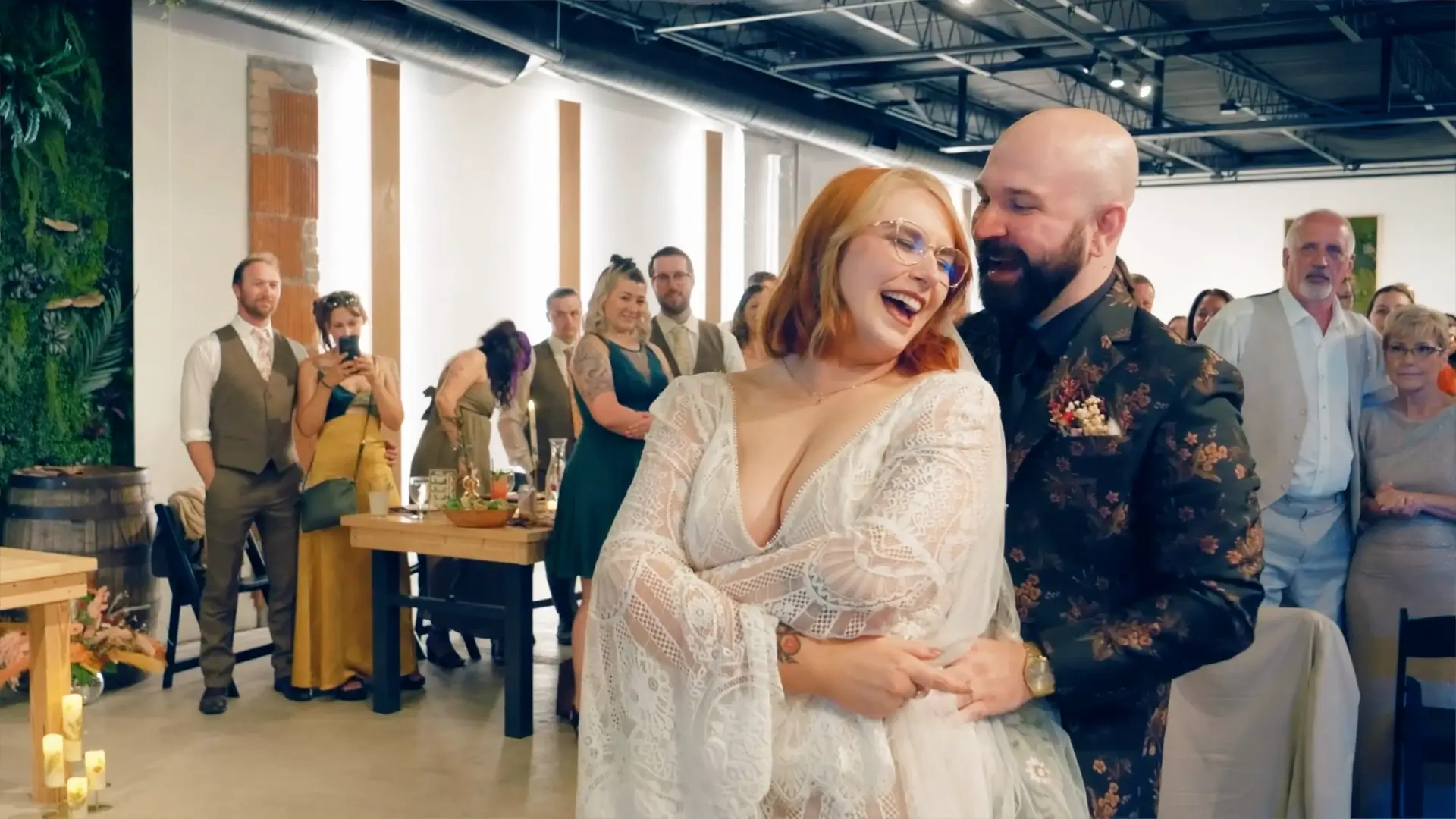 Kate and Zachery share a joyful moment during their wedding reception at Solarium by Watermark Brewing Company. The bride wears a lace dress, and both are smiling amidst a lively crowd of guests.