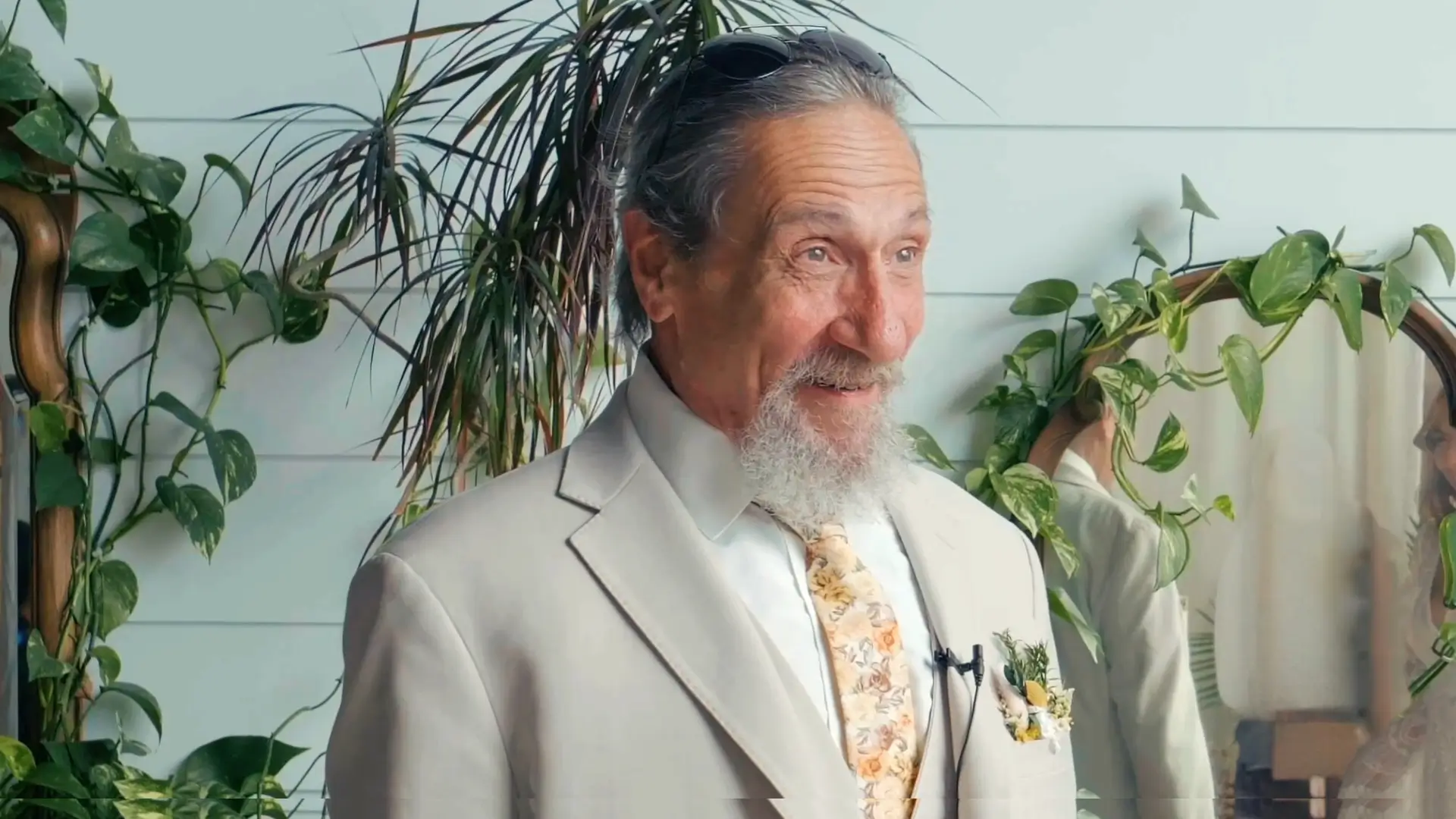 A smiling man with a grey beard and sunglasses – Kate's Father – wears a light beige suit with a floral tie, standing near indoor plants and a round mirror, with an excited expression on his face as he sees his daughter's dress for the first time.
