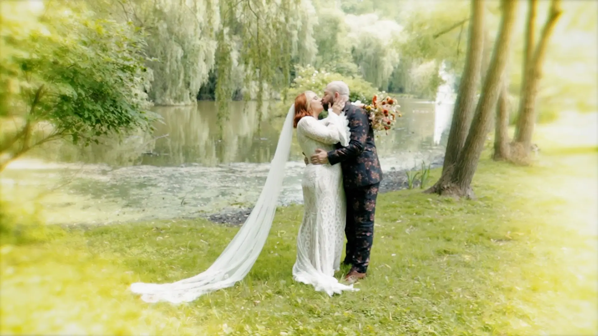 Kate and Zack share a kiss by a serene pond in Friendship Botanic Garden, beautifully captured in this wedding video. Kate wears a flowing white gown, while Zack dons a patterned suit, surrounded by lush greenery that reflects their artsy vibe.