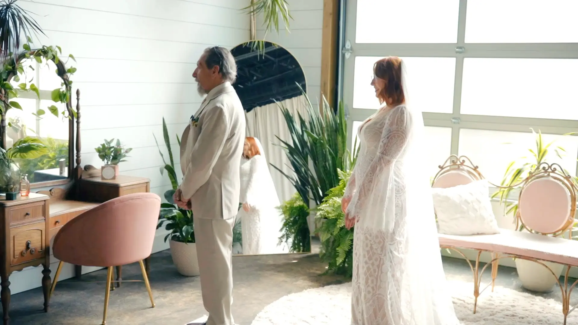 Bride in a lace gown smiles at an older man in a beige suit inside a sunlit room with plants and vintage furniture. The scene is likely set in a bridal suite at the Solarium by Watermark Brewing Company, capturing an intimate moment before the ceremony.