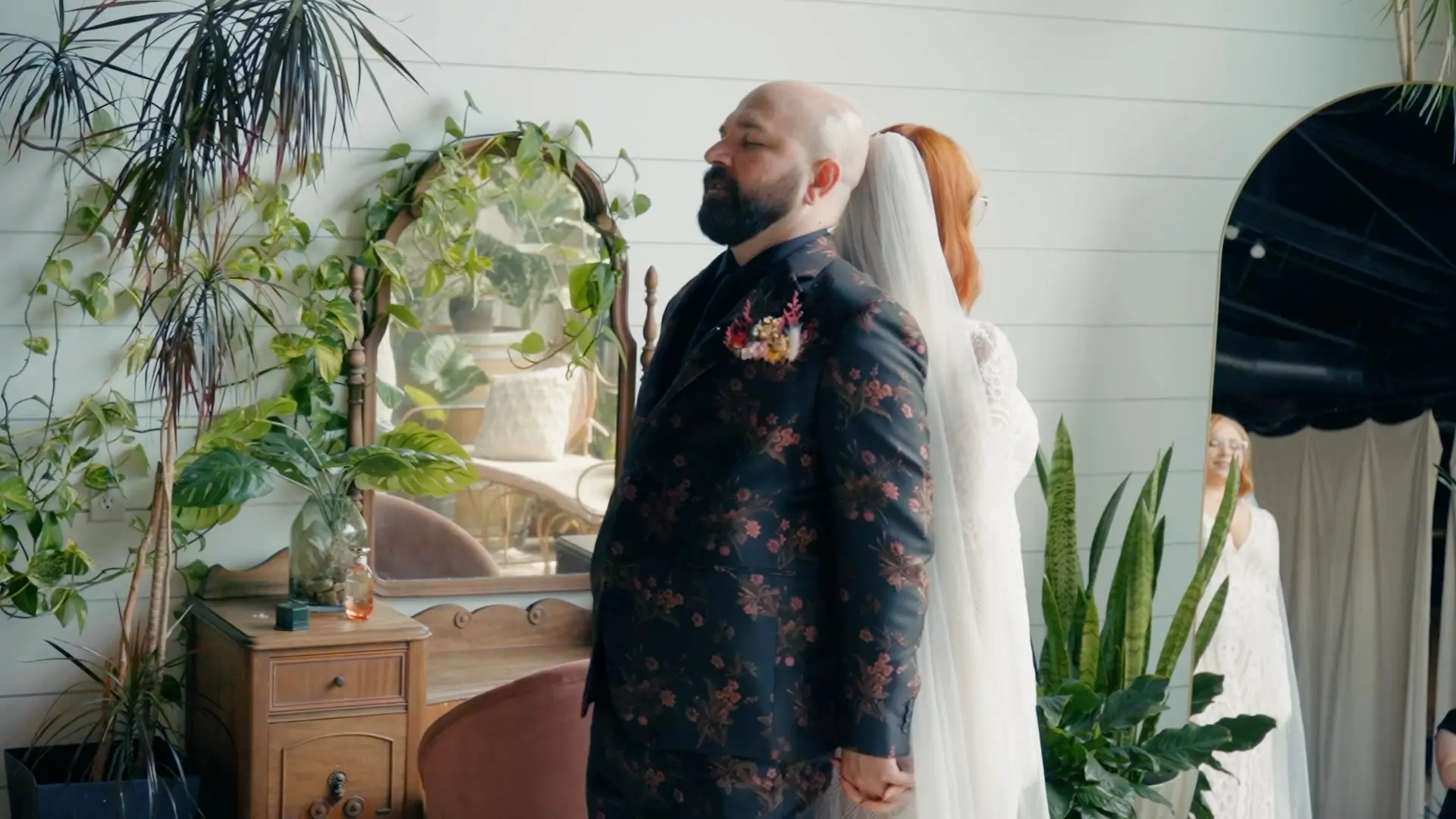Bride and groom sharing a tender first look moment, standing back-to-back in an airy, plant-filled room. The groom wears a floral suit while the bride smiles, reflected in a mirror. Lush Michigan greenery surrounds them, enhancing the warm atmosphere.