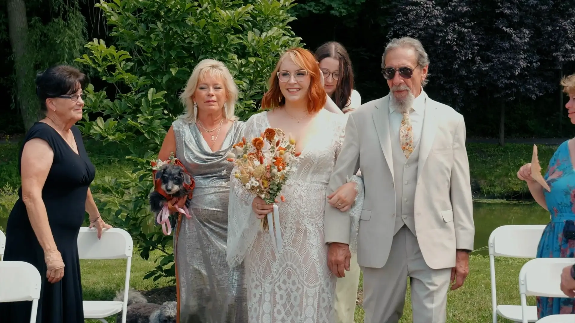 Bride Kate walks down the aisle at Friendship Botanic Garden in Michigan City, Indiana, with her family. She is wearing a lace gown and holding a bouquet of earthy flowers. Lush greenery surrounds the scene on a sunny day.