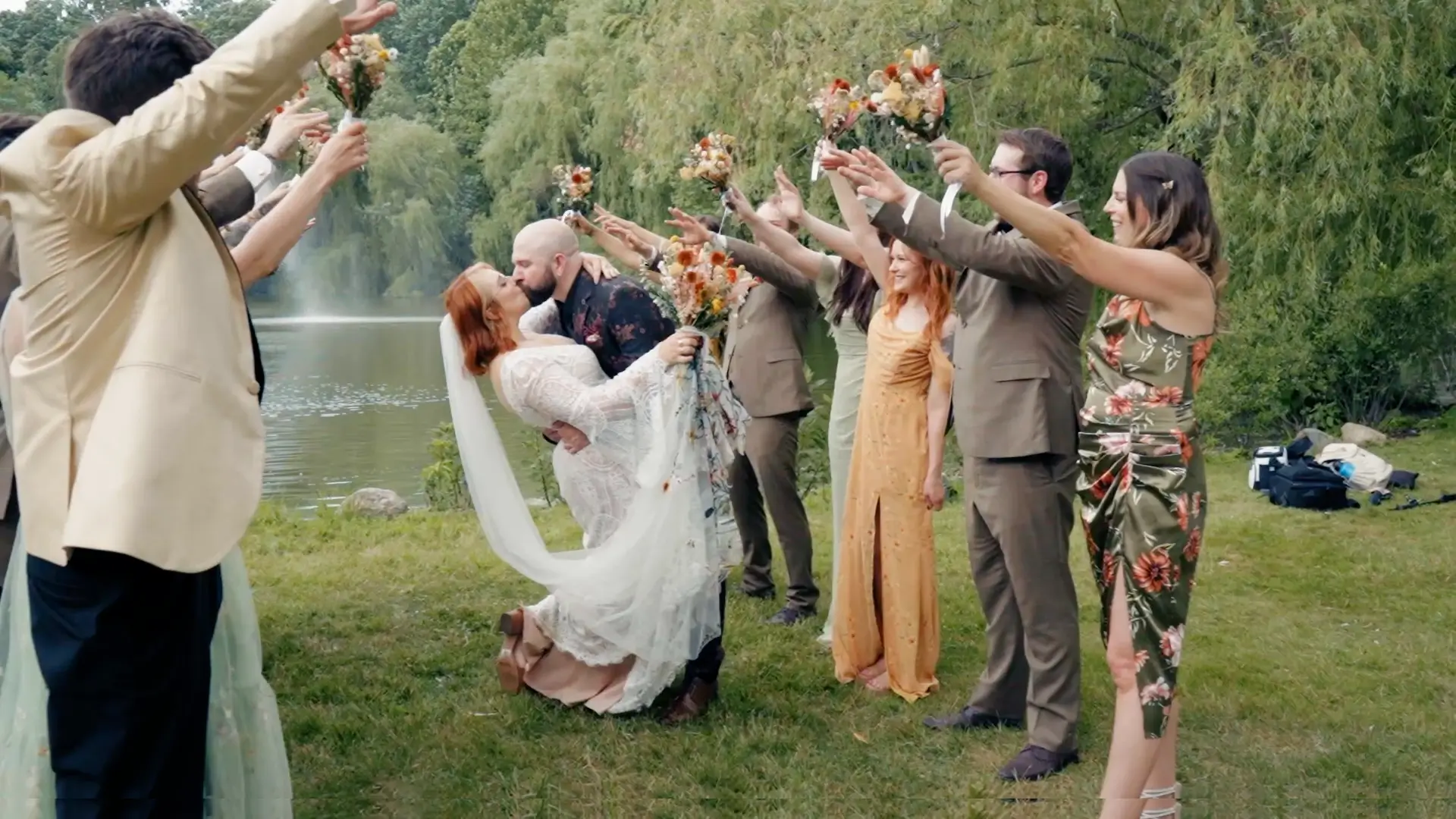 Bride and groom, Kate and Zack, are kissing in a dip pose by a lake, surrounded by their wedding party holding bouquets aloft at Friendship Botanic Garden, Michigan City, Indiana. The scene is set outdoors with trees and greenery in the background.