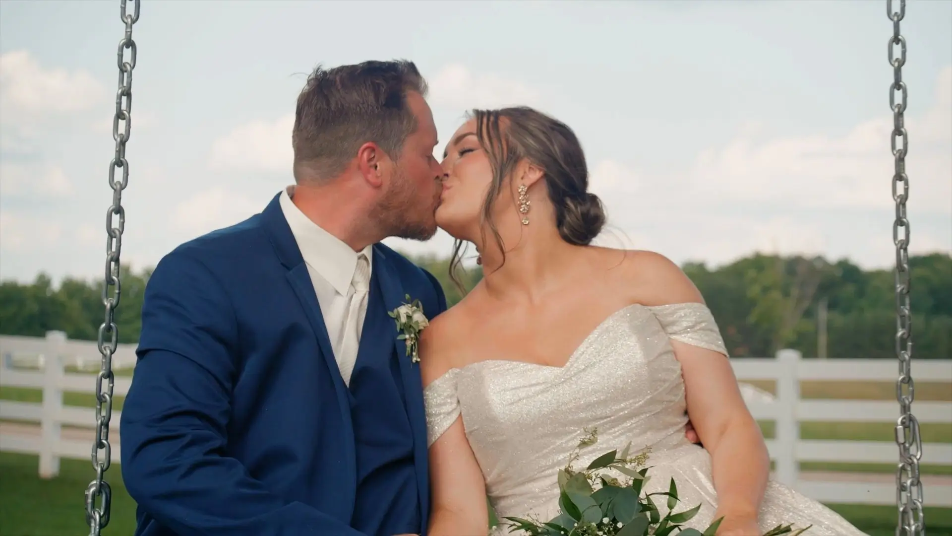 Bride Brittany in an off-shoulder white gown and groom Dakota in a blue suit kiss while sitting on a swing at Sonshine Barn Wedding and Event Center in Gaylord, Michigan, on their wedding day, July 13, 2024. The bride holds a bouquet.