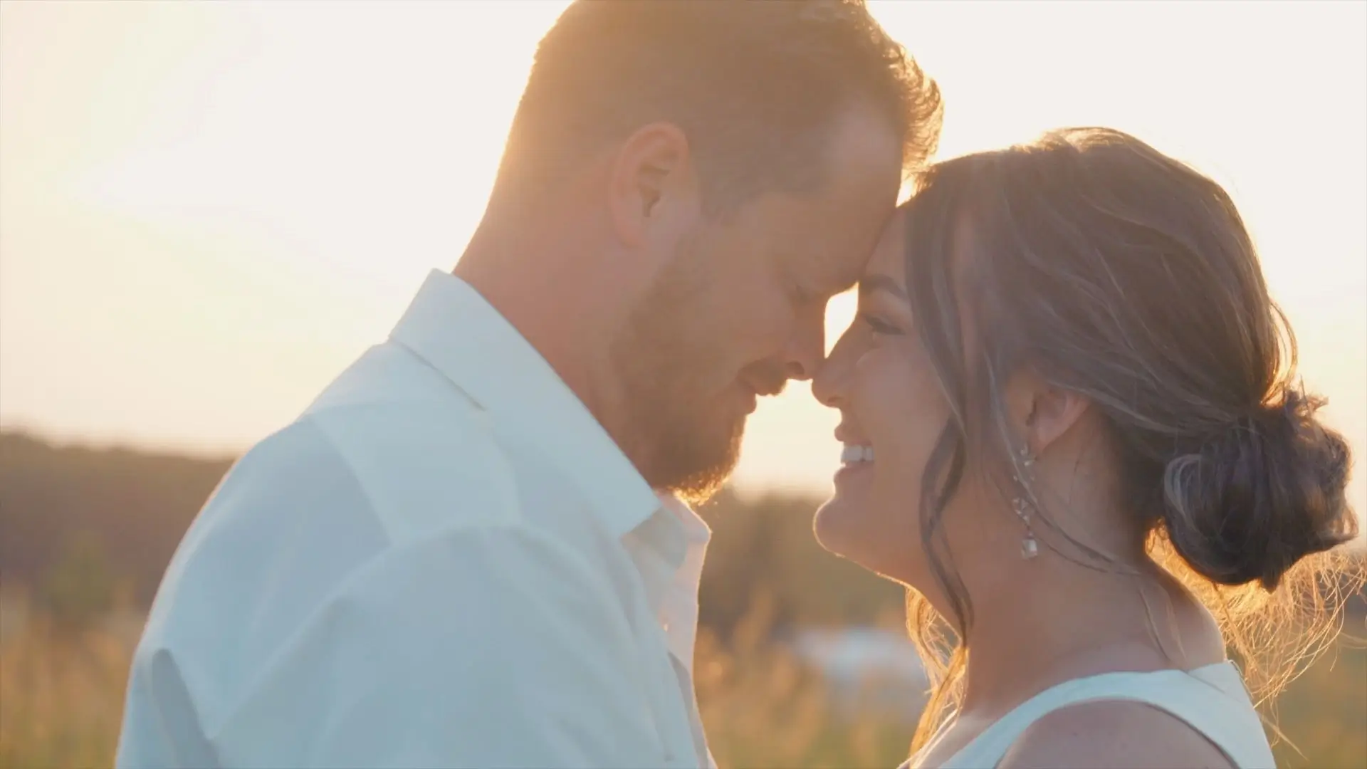 Brittany and Dakota smile with their foreheads touching as the sun sets behind them at Sonshine Barn Wedding and Event Center in Gaylord on July 13, 2024. Brittany wears her hair up and shows off dangling earrings, while Dakota wears a light shirt.