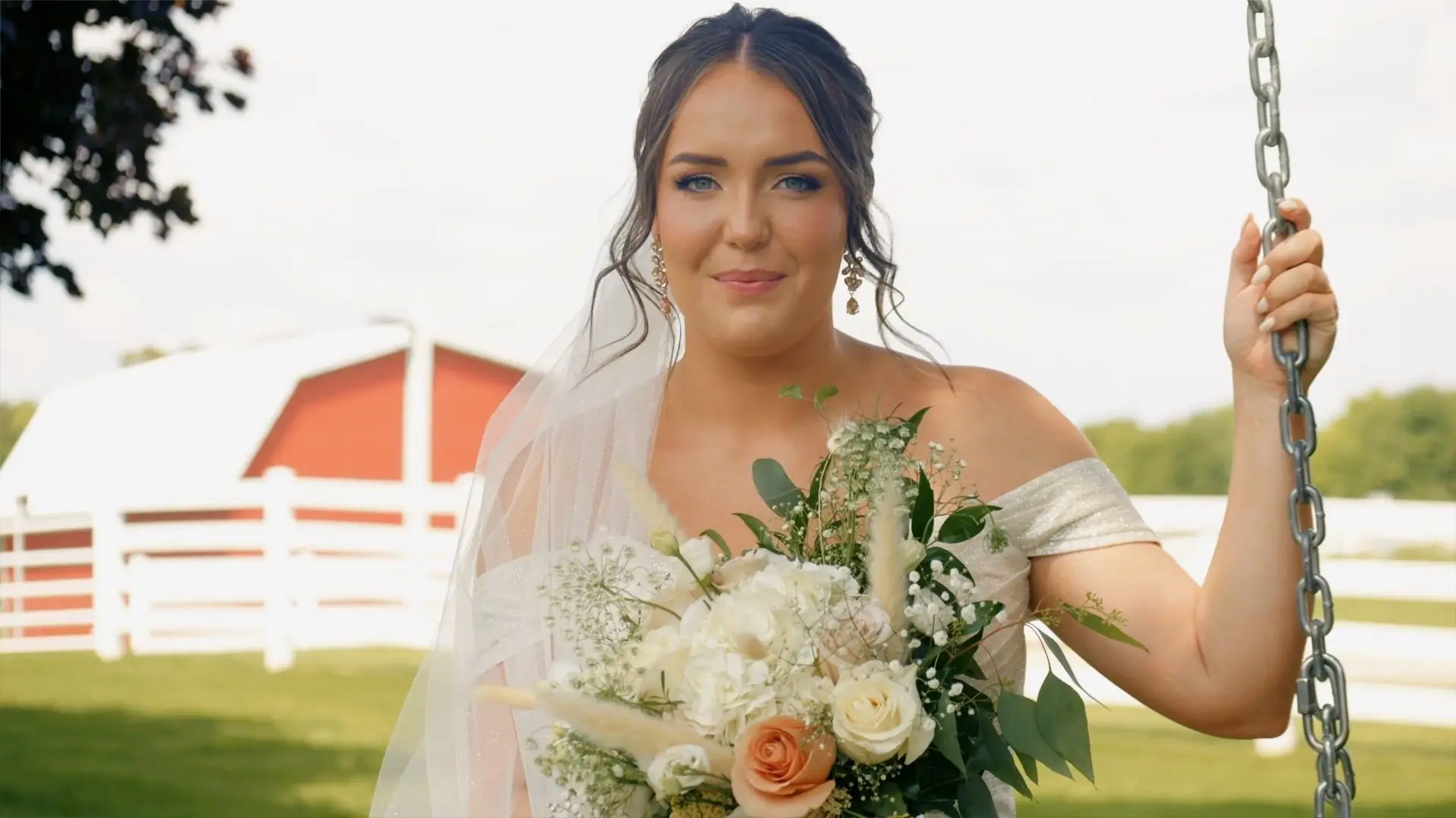 Bride Brittany holding a bouquet and sitting on a swing at Sonshine Barn Wedding and Event Center in Gaylord, Michigan. She is dressed in her wedding gown with a red barn and white fence visible in the background.