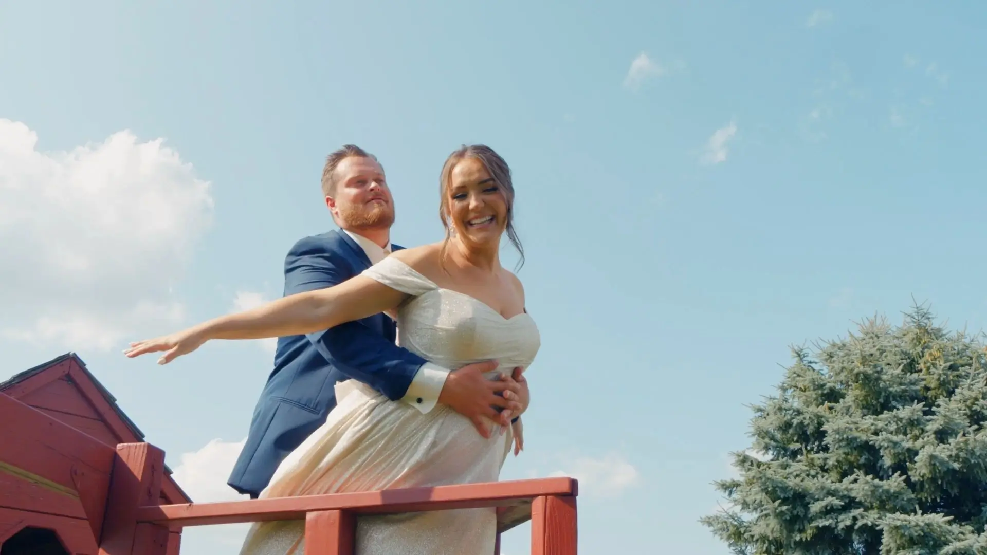 Bride Brittany and groom Dakota pose playfully like the iconic scene from "Titanic." They stand on a red wooden structure with a blue sky and a green tree behind them at Sonshine Barn Wedding and Event Center in Michigan.