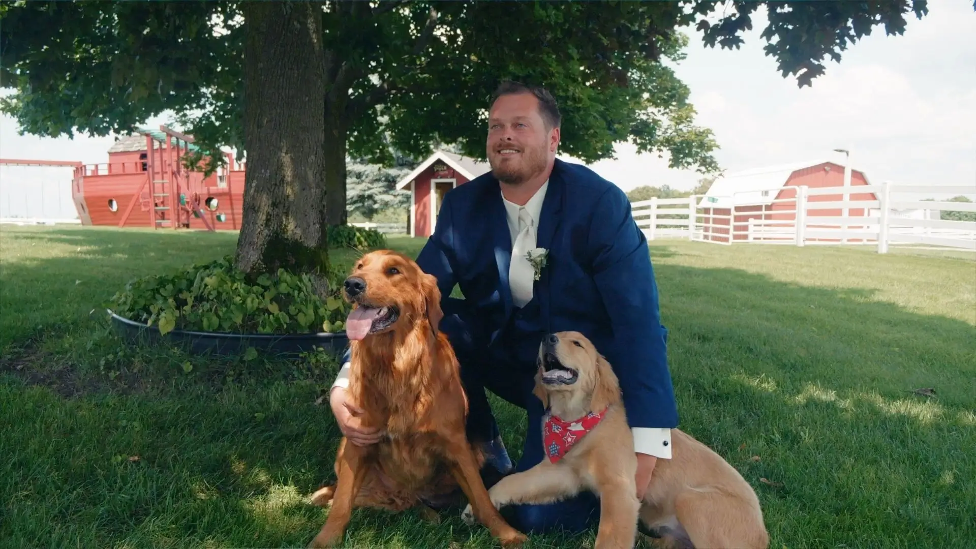 In this snapshot from their wedding video, Dakota, dressed in a navy suit, poses under a tree with two happy dogs at the Sonshine Barn Wedding and Event Center in Gaylord, Michigan. Red barns and green lawns provide a picturesque background.