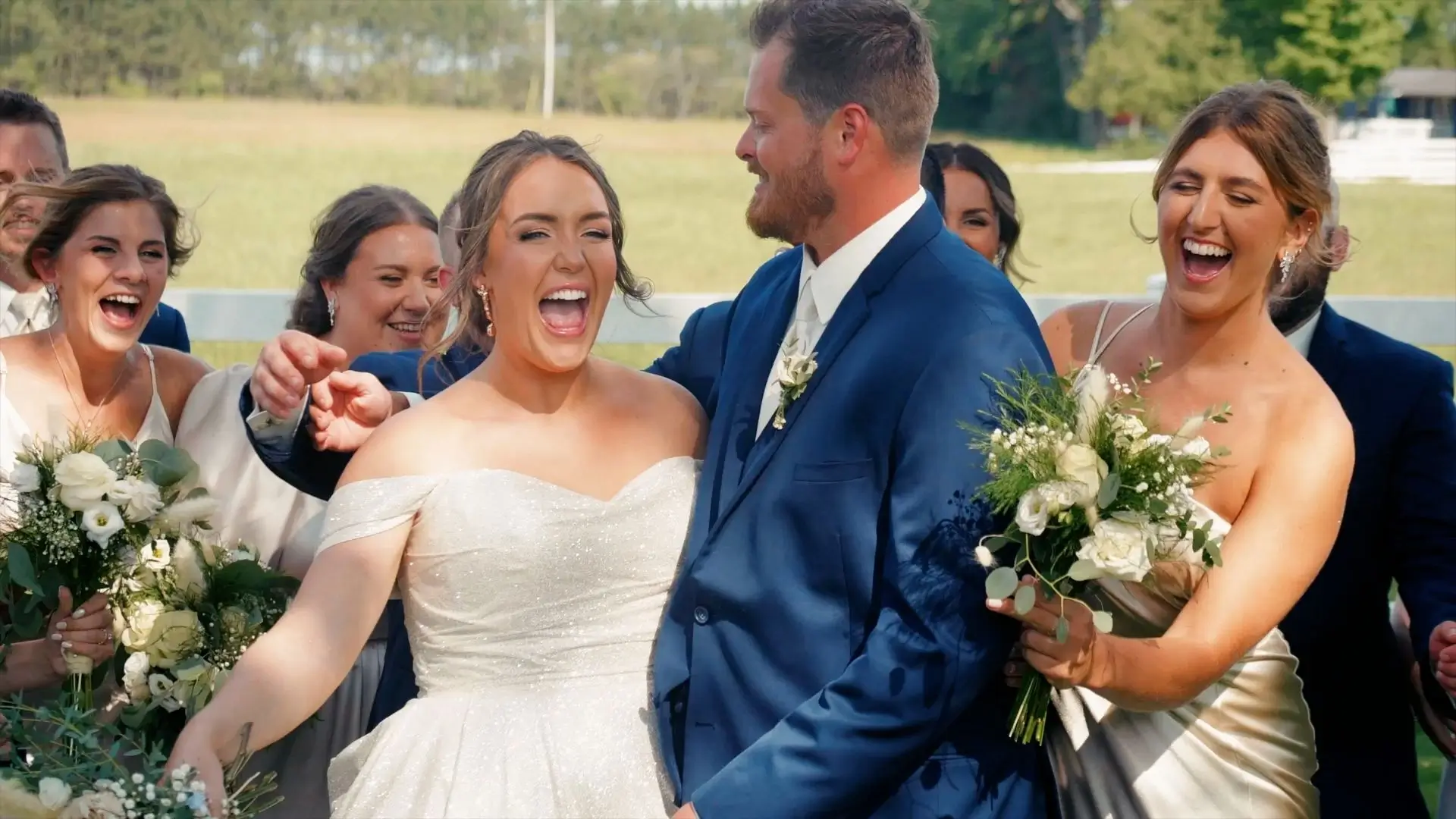 Bride Brittany and groom Dakota smiling joyfully, surrounded by their laughing wedding party at the Sonshine Barn Wedding and Event Center in Gaylord. The bride is in a white dress, and the groom wears a blue suit. Bridesmaids hold white floral bouquets.